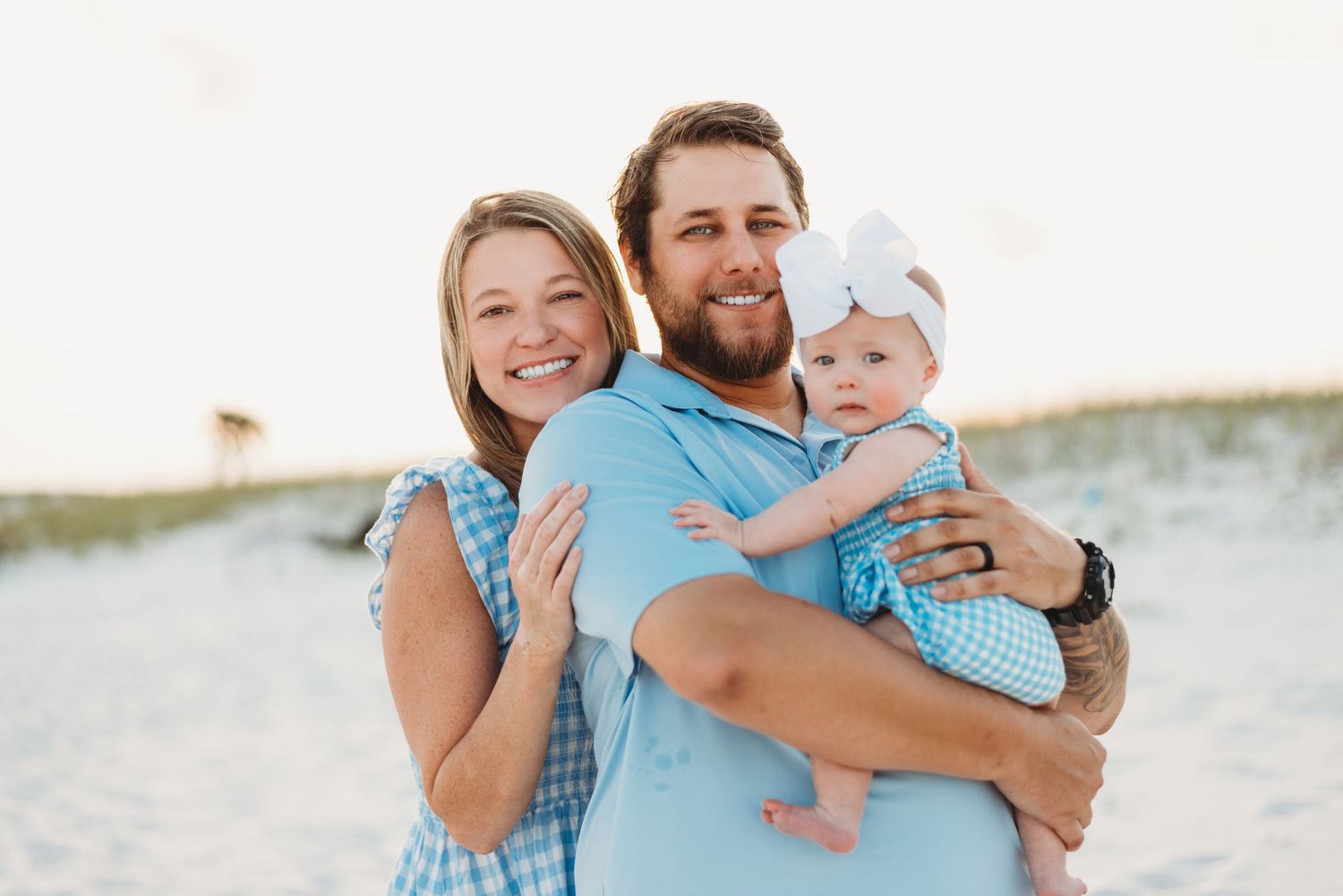 Smiling couple holding their baby girl in a blue bow on a bright sandy beach.