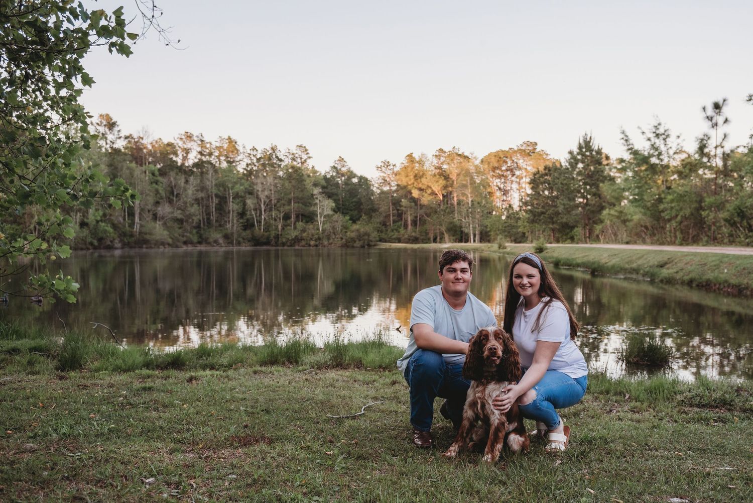Smiling couple posing with their brown dog by a calm reflective pond at sunset.