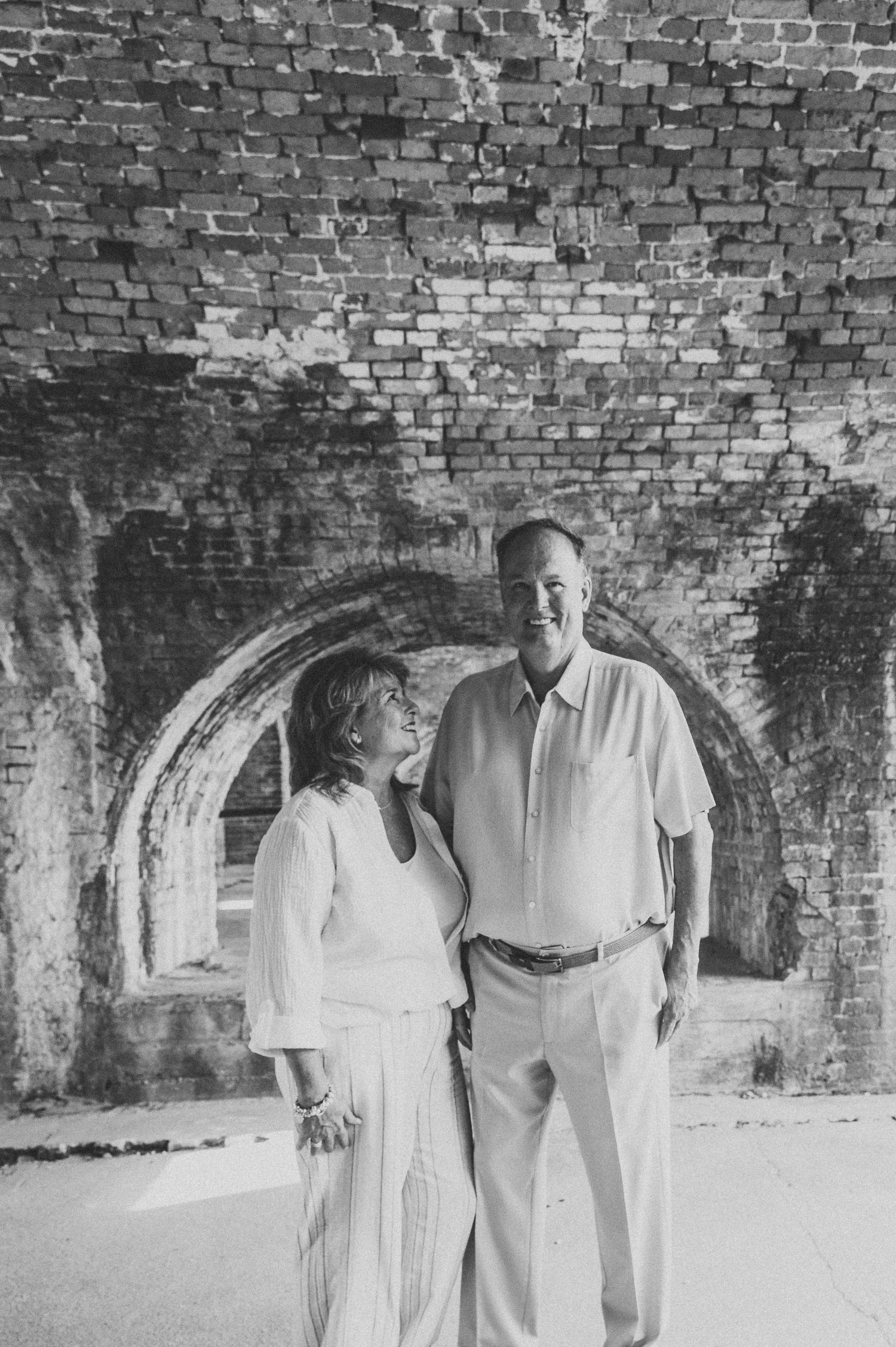 Black and white photo of a smiling couple standing together under historic brick arches.