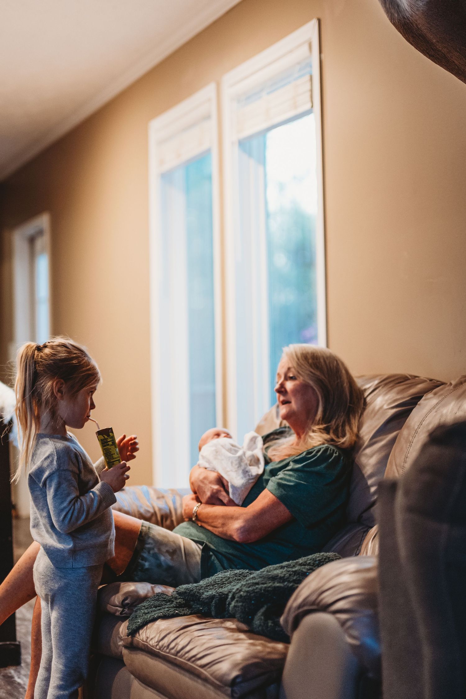 Grandmother sitting on couch holding newborn while young granddaughter stands beside her.