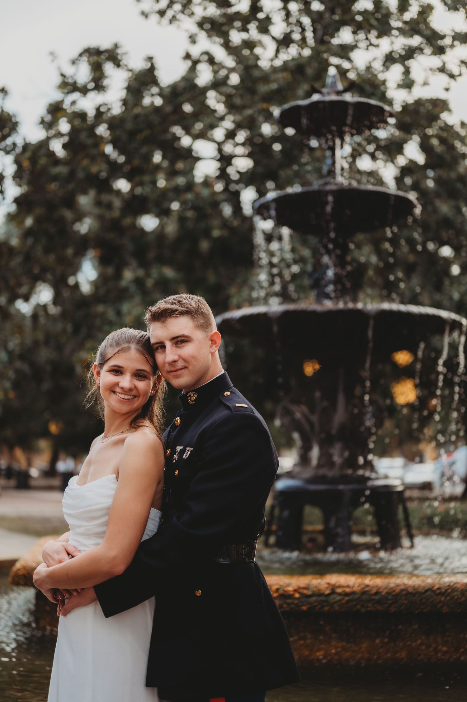 Couple in wedding attire poses by ornate fountain, groom in Marine dress uniform, bride in white gown.