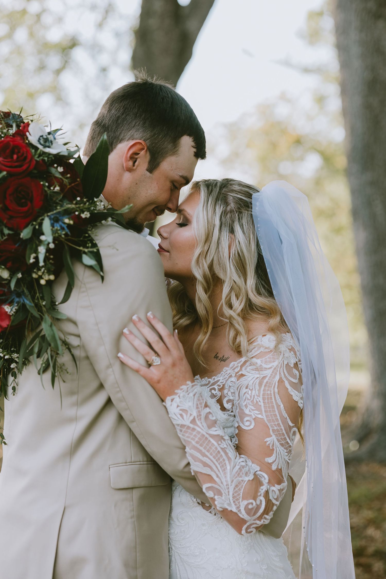 Bride and groom share a romantic forehead touch, bride in lace gown with veil holding red roses bouquet outdoors.