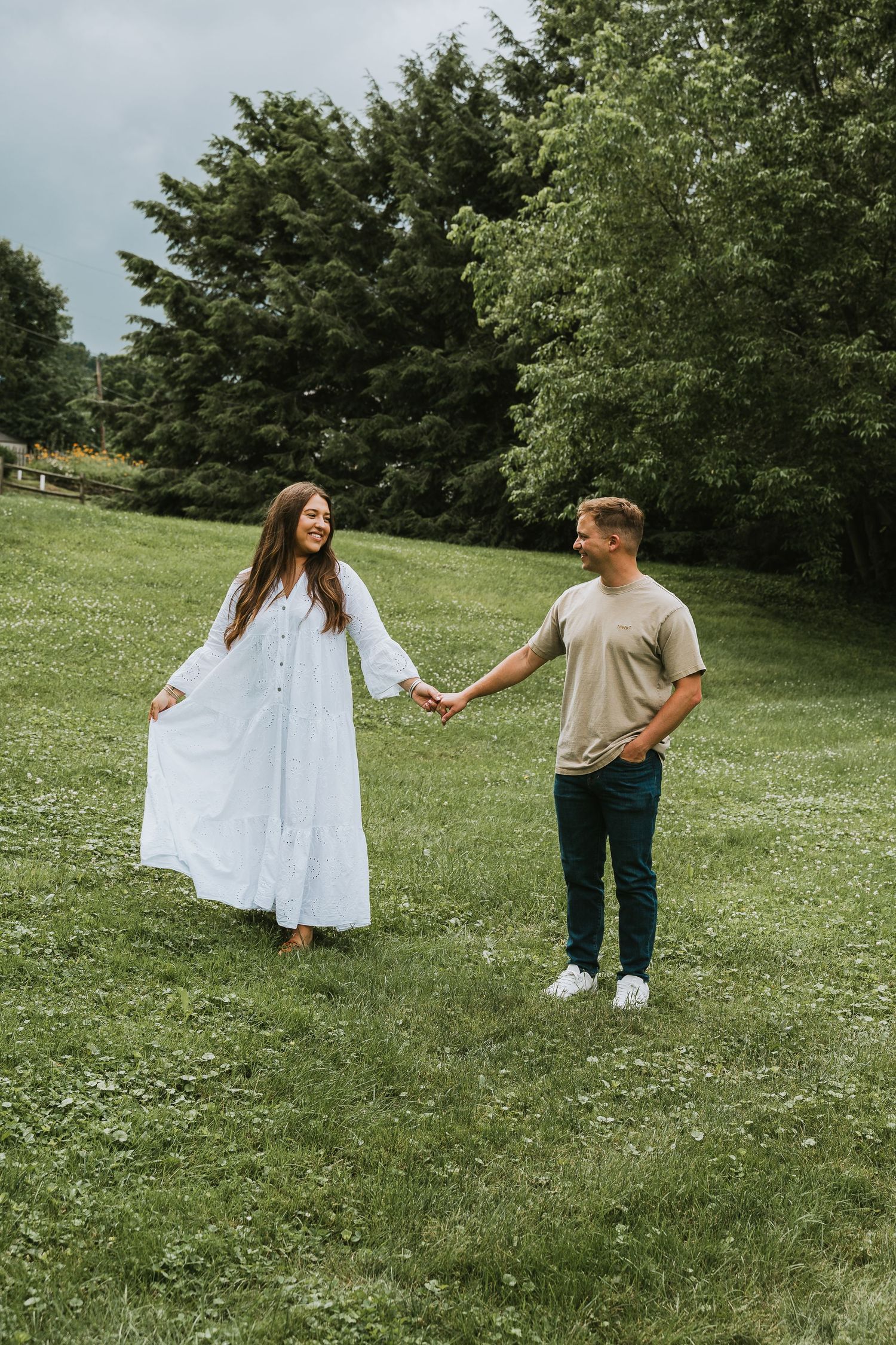 A couple holds hands on a green meadow, woman in white dress, man in tan shirt, with trees in background.