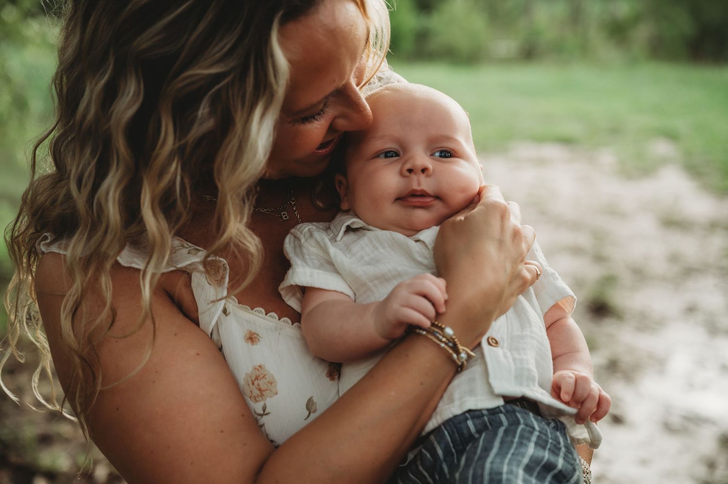 A loving mother with blonde wavy hair cuddles her smiling baby outdoors in a serene natural setting.
