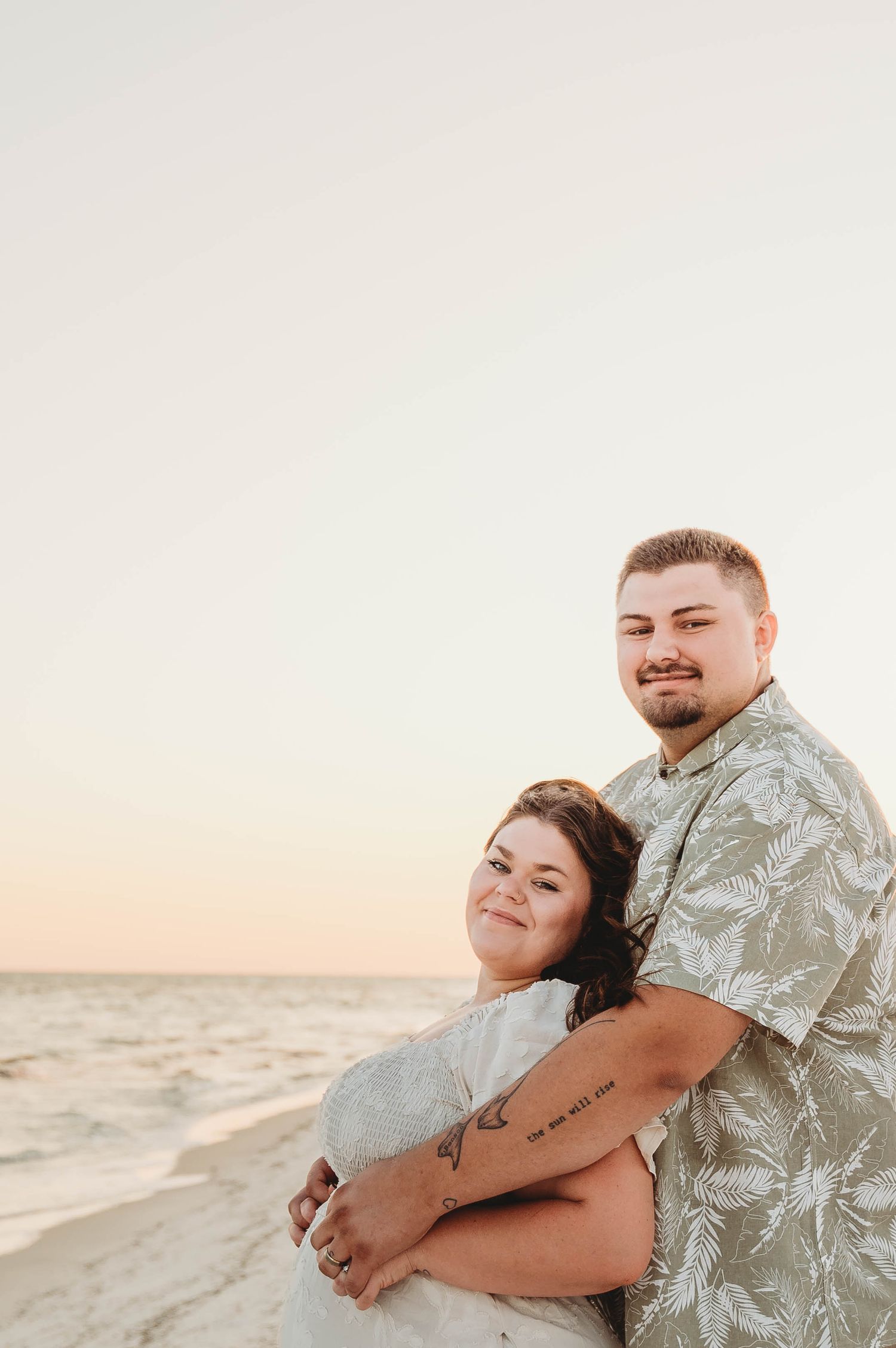 A couple embraces warmly on a beach at sunset, smiling at the camera.