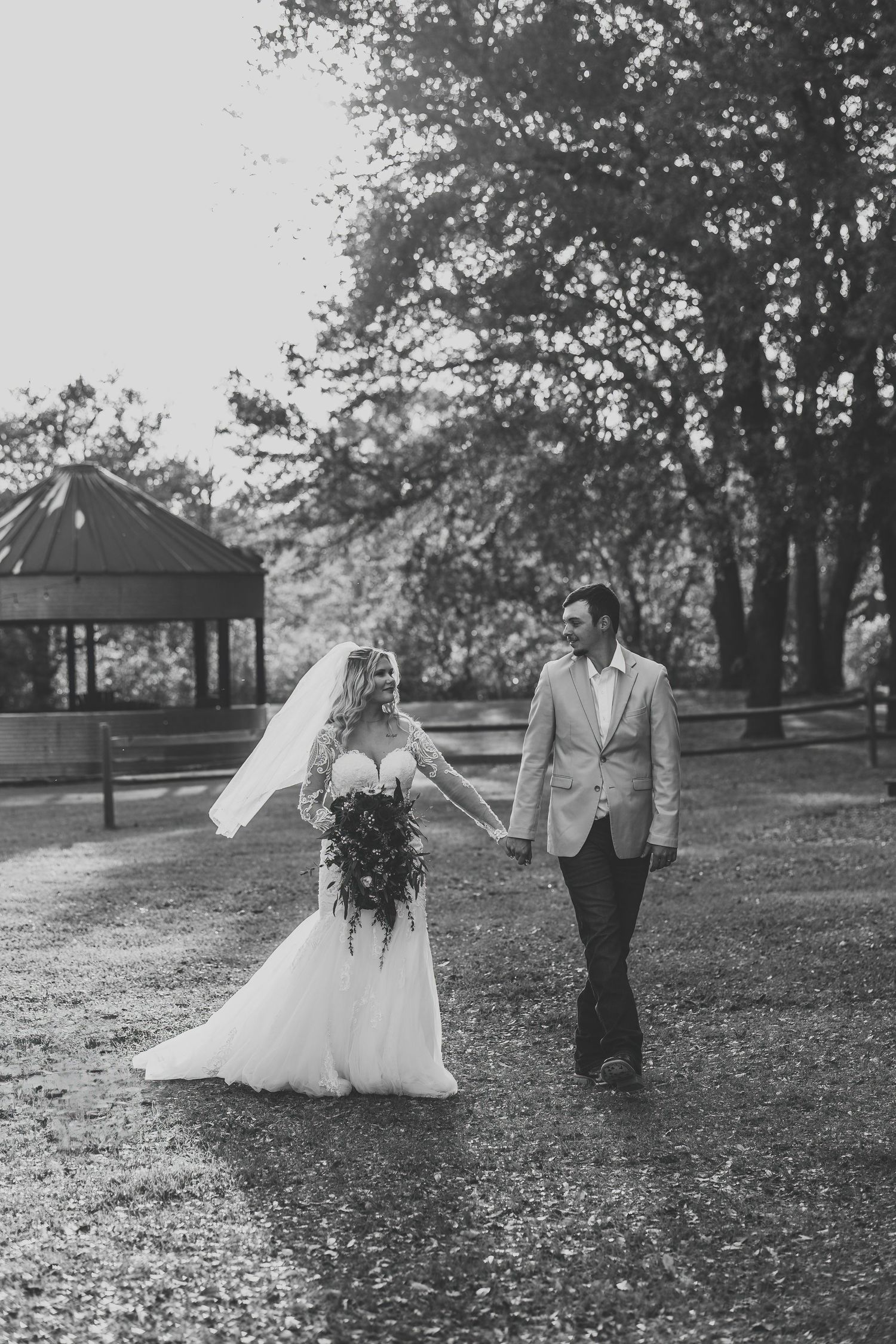 Black and white photo of bride and groom walking hand in hand outdoors, veil blowing in wind.