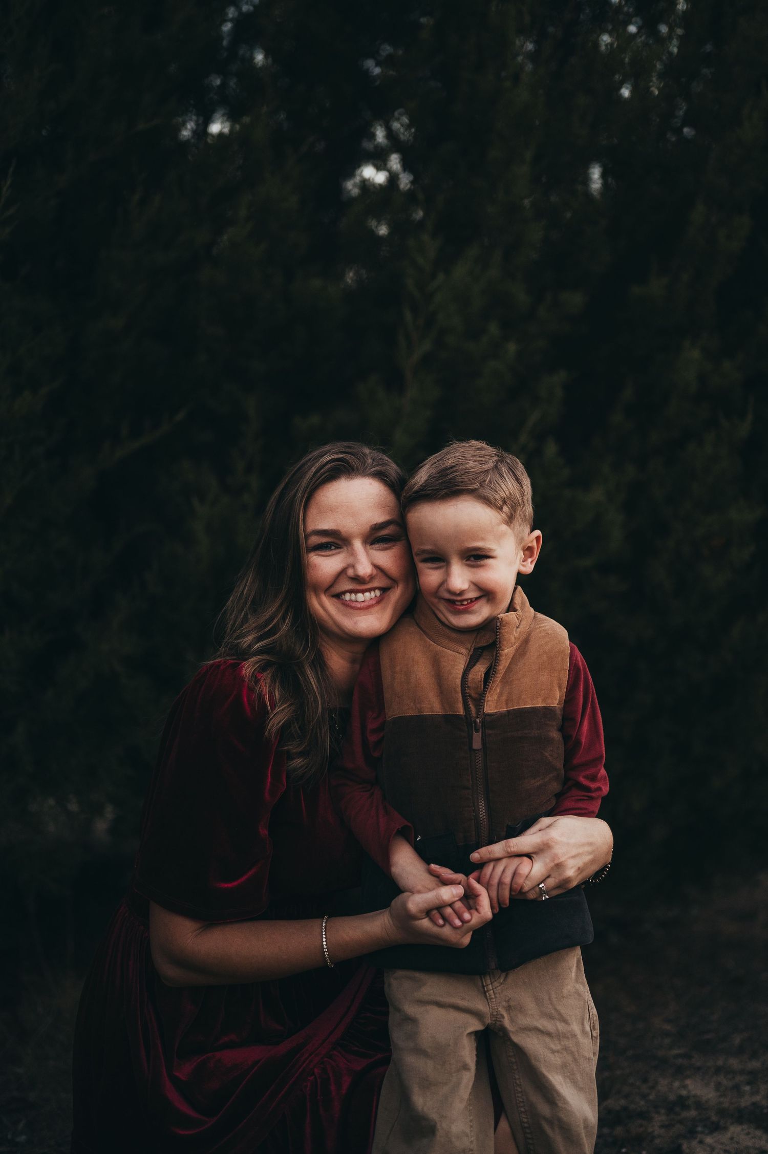 A smiling woman in a red dress hugs a young boy in a brown vest outdoors near evergreen trees.