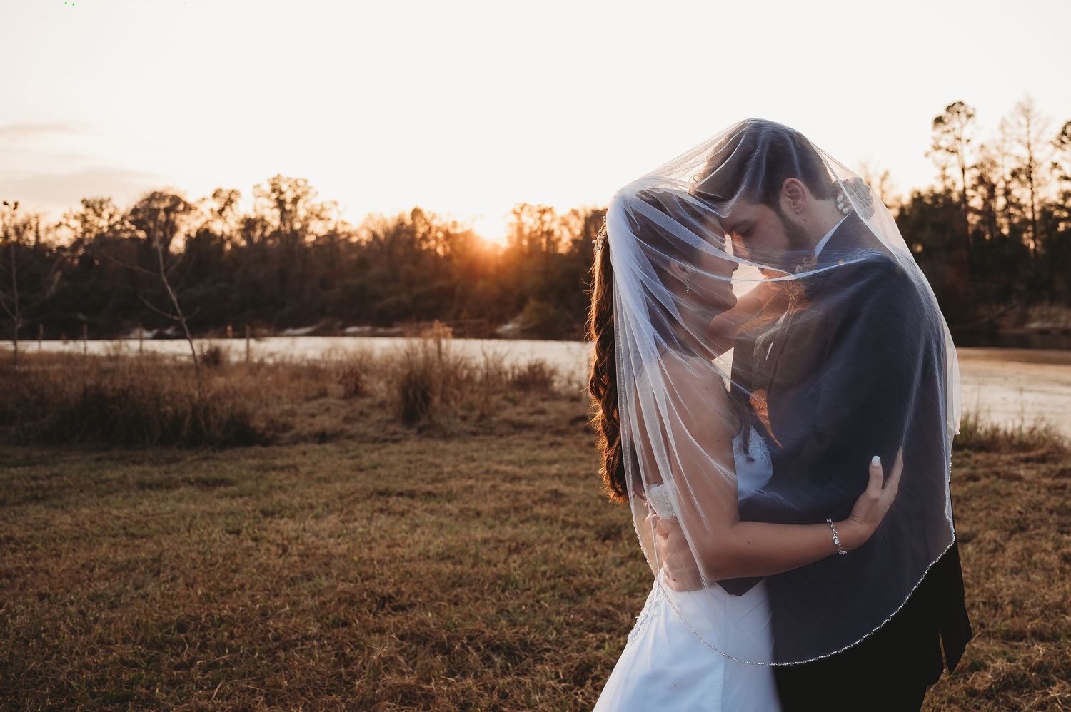 Bride and groom sharing a romantic kiss under a veil at sunset by a serene lake.