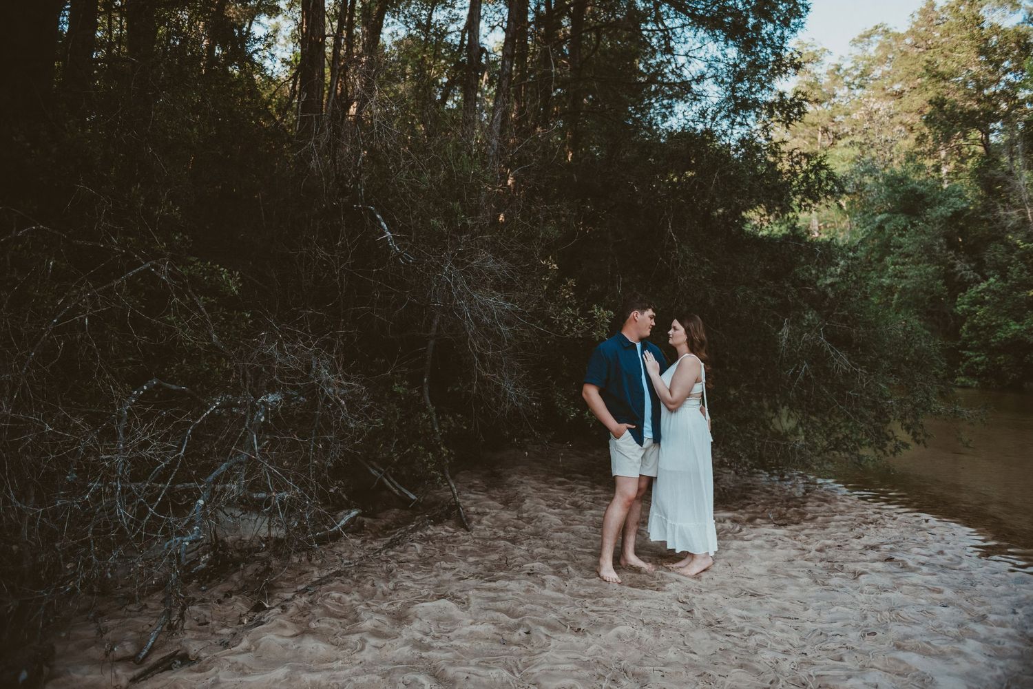 Engaged couple standing barefoot on sandy ground surrounded by lush green trees during golden hour.