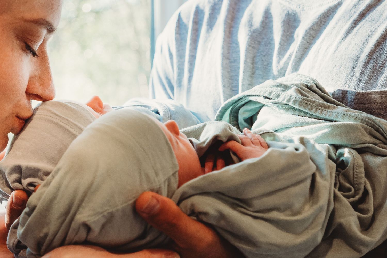 A loving mother cradles and kisses her newborn baby wrapped in a mint green blanket.