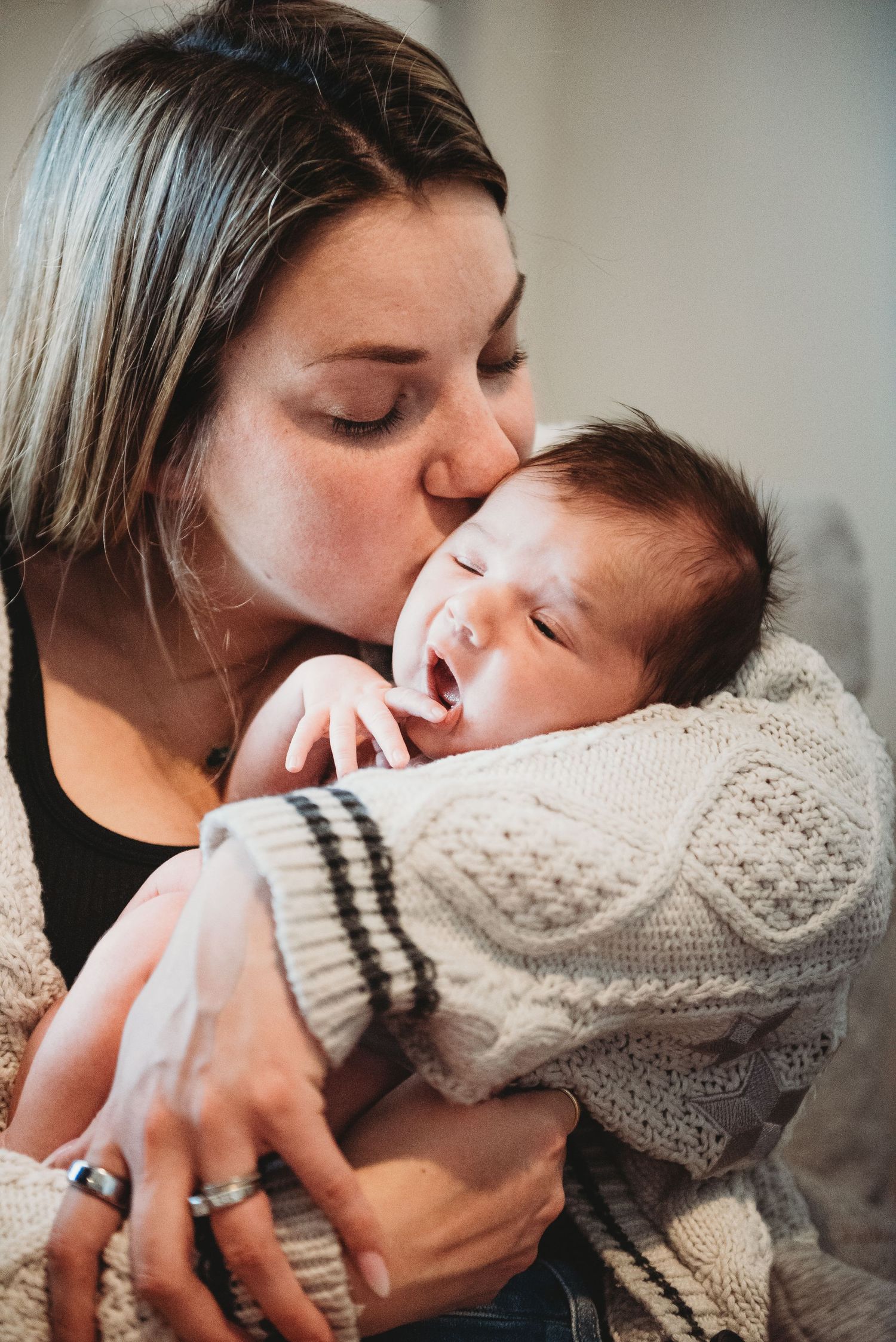 Mother tenderly kissing her newborn baby wrapped in a knit sweater.