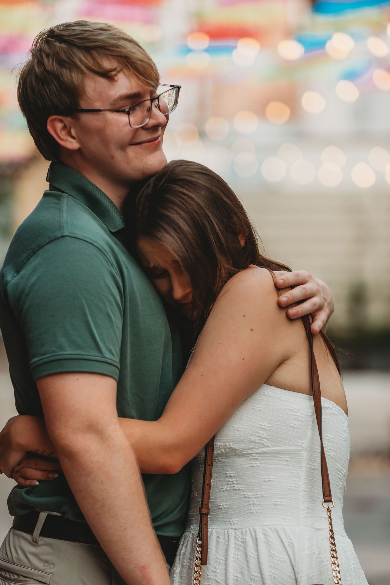 A young couple embracing at a fair, man in green polo and glasses hugging woman in white dress.