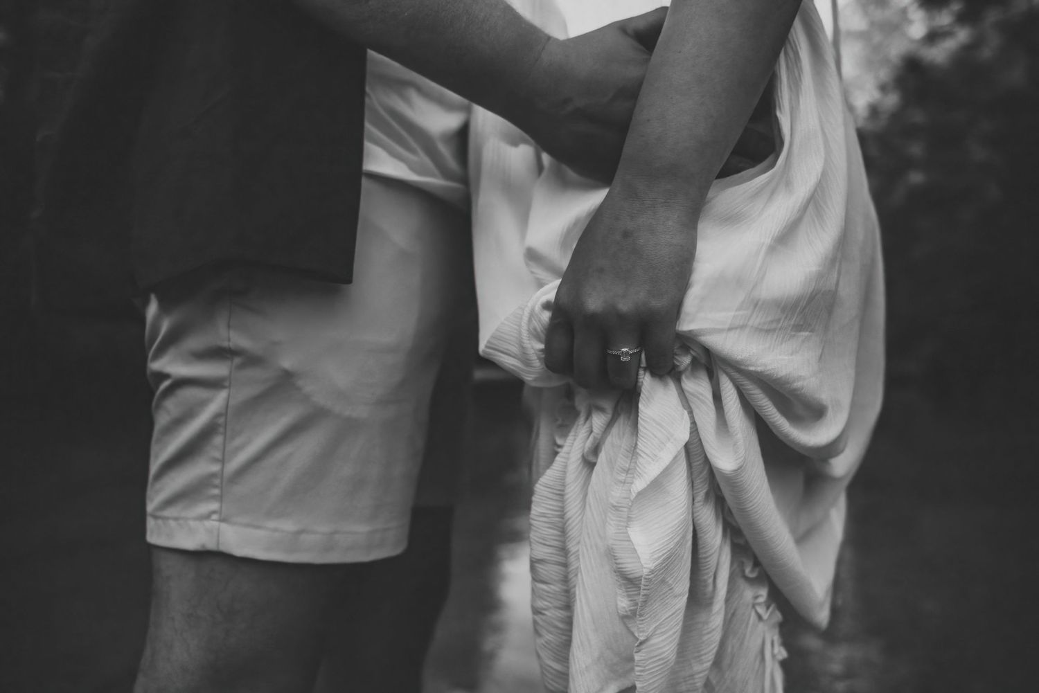 Close-up black and white shot of couple holding hands, woman wearing engagement ring and white dress.