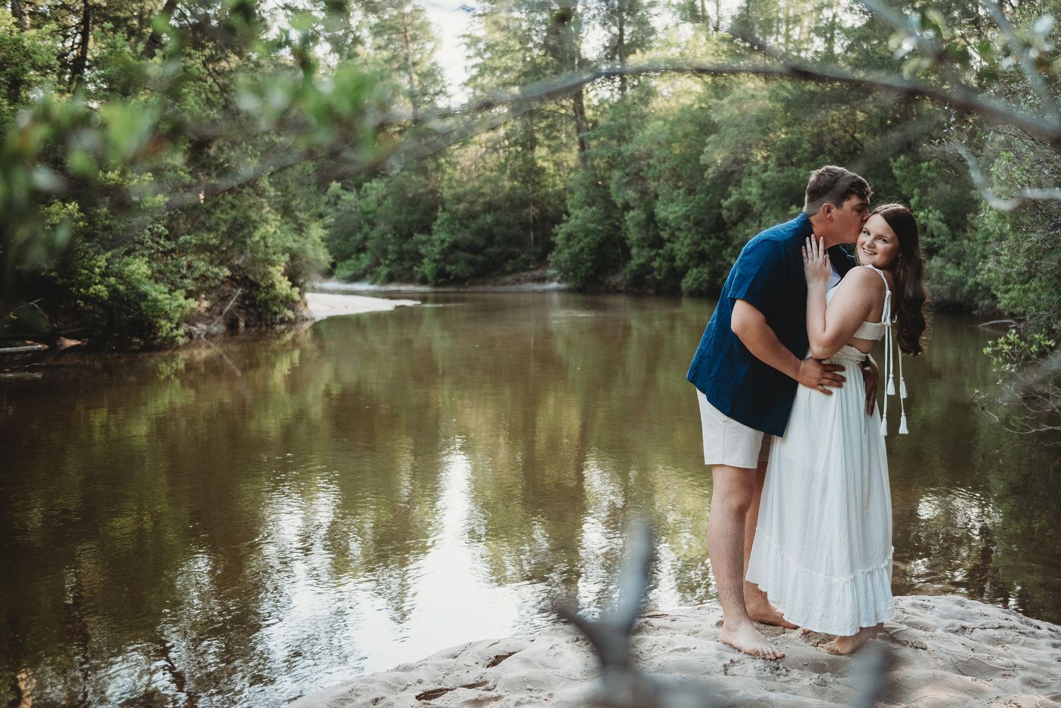 Couple sharing a kiss while standing on rocks beside a calm reflective river in the forest.