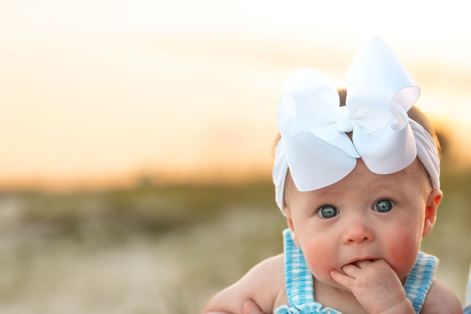 Close-up portrait of a cute baby girl with a large white bow headband at sunset.