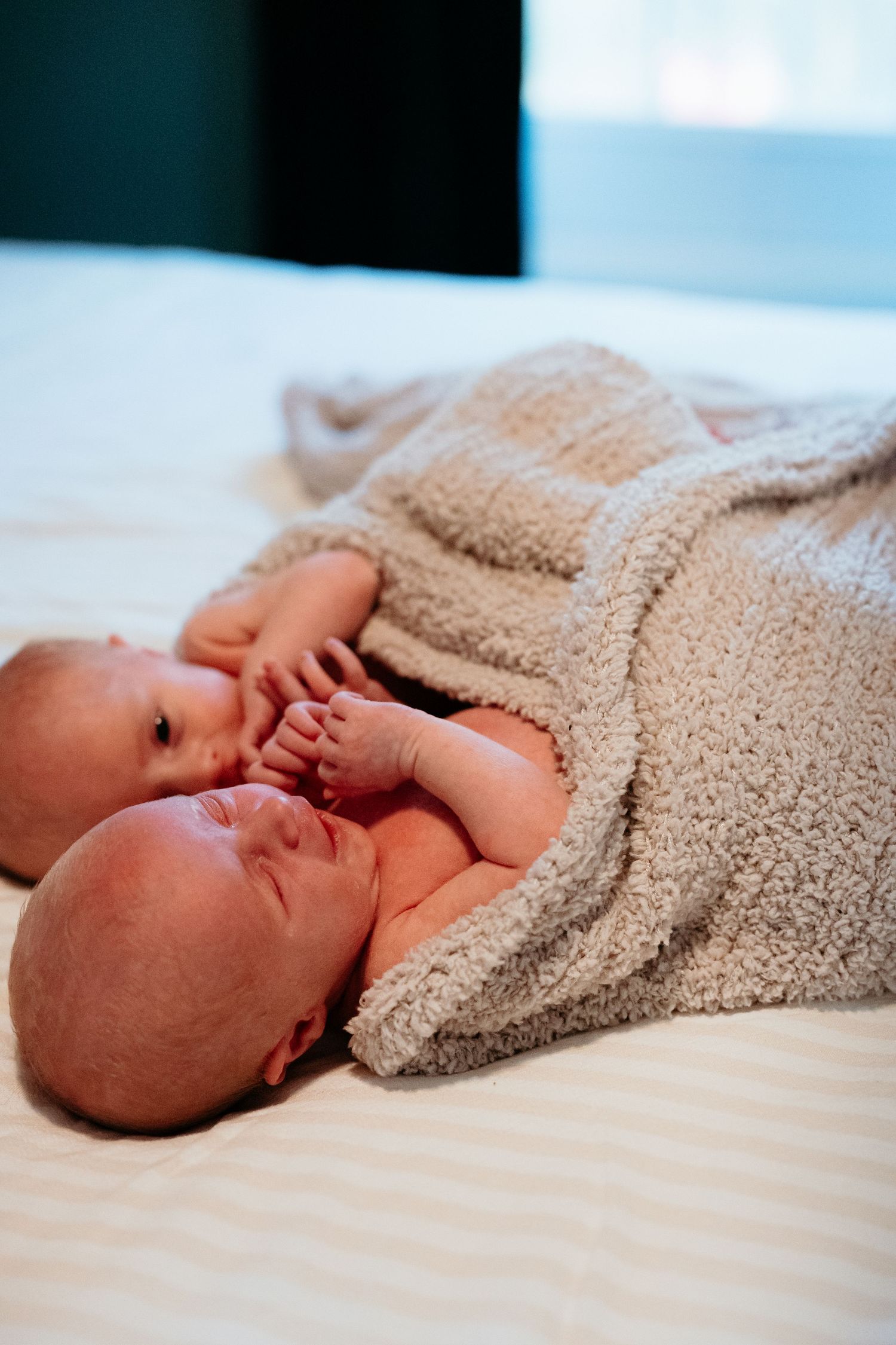 Two newborn twins lie side by side wrapped together in a soft beige blanket on white bedding.
