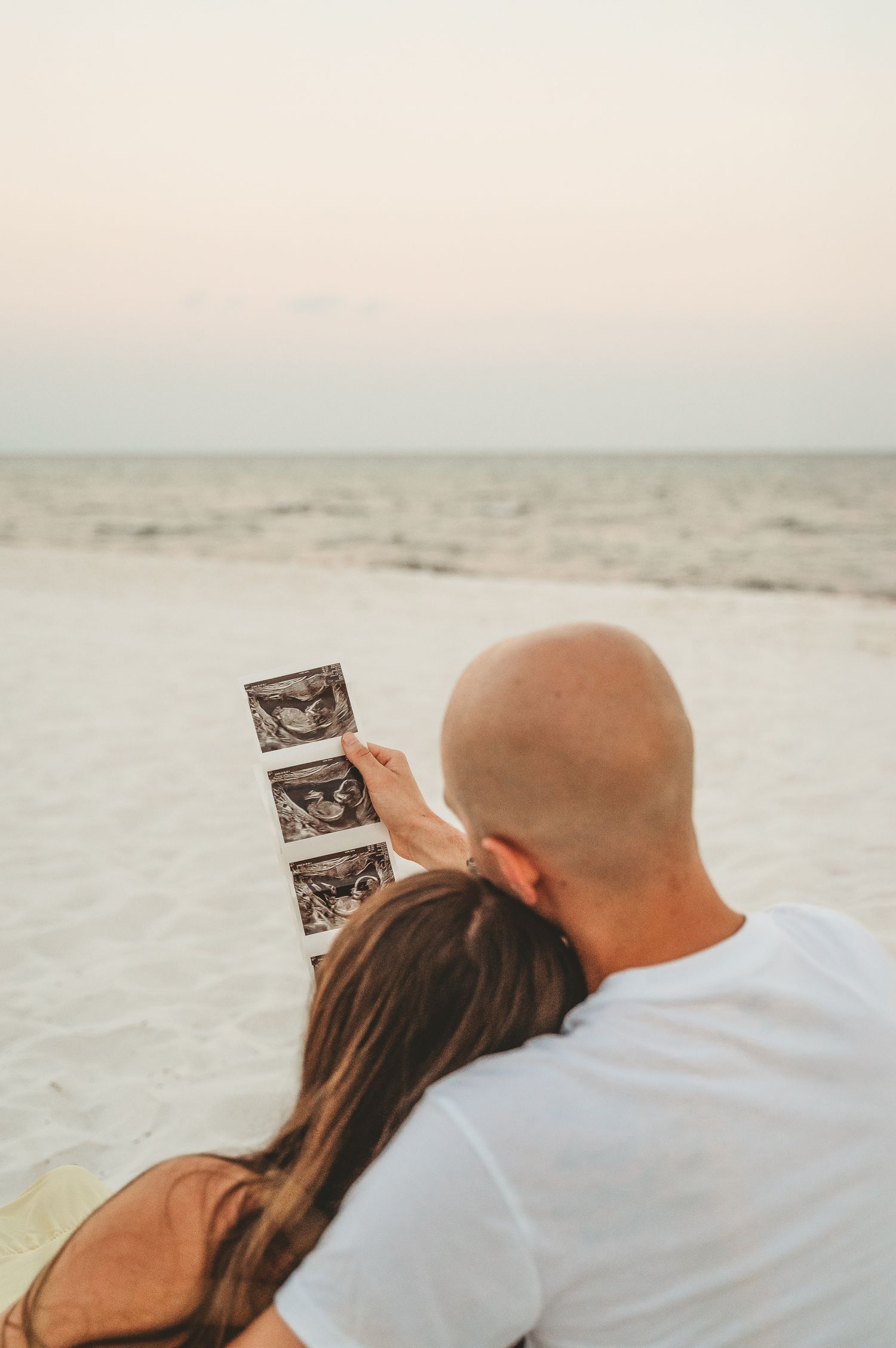 Couple sitting together on a beach at dusk looking at ultrasound photos to announce their pregnancy.