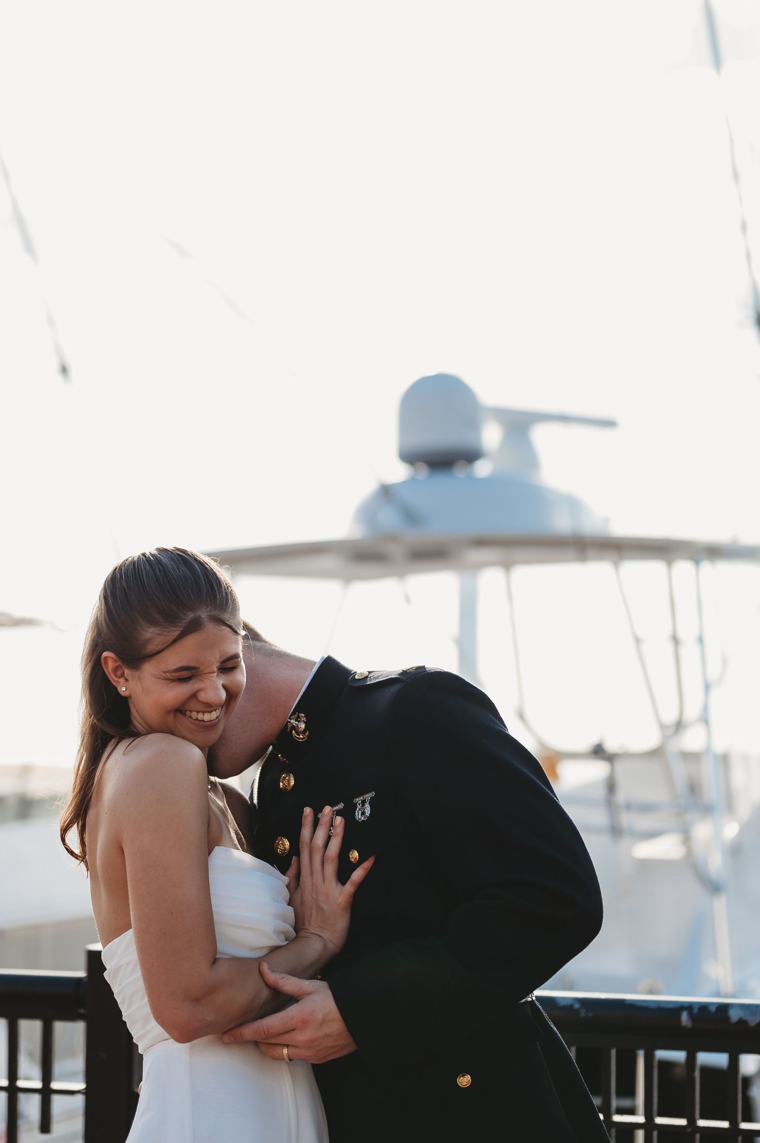 A smiling bride in a white dress embraces a groom in a military uniform near a marina.