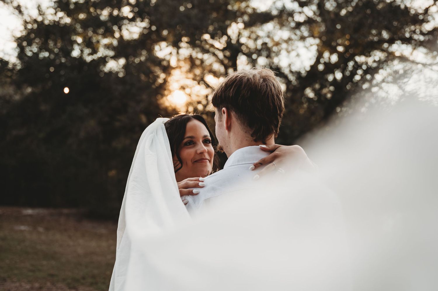 A bride and groom embrace under a flowing white veil at golden hour, surrounded by lush trees.