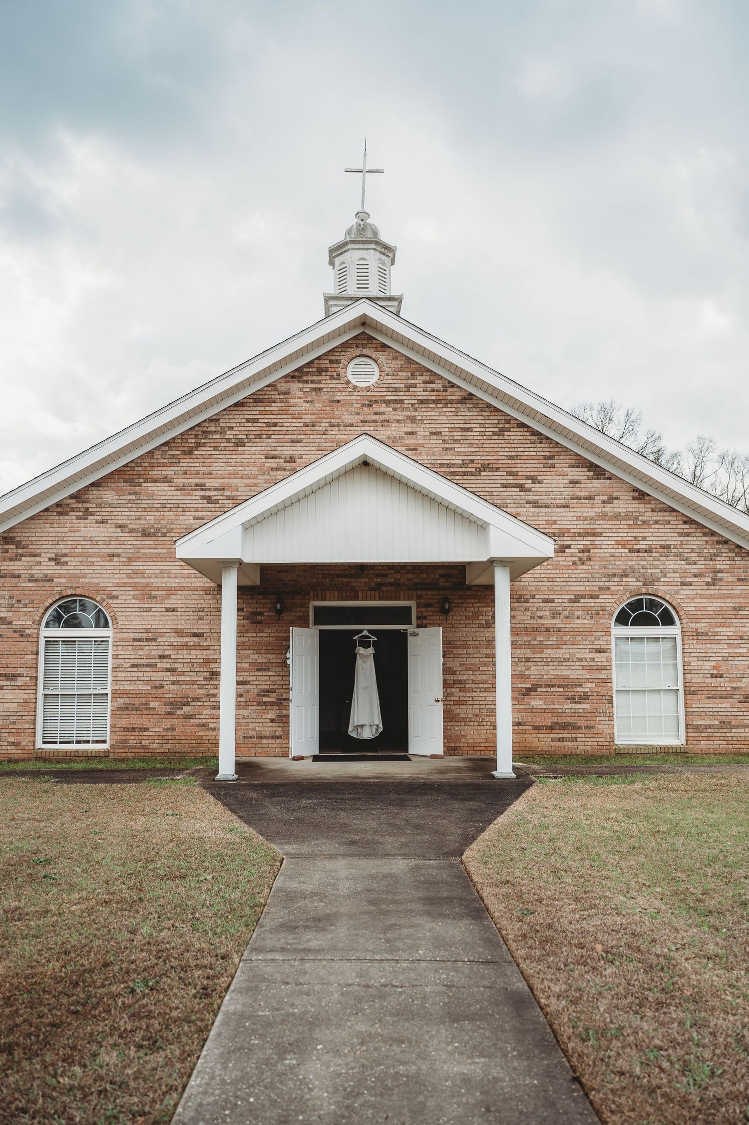 A white wedding dress hangs on the door of a charming brick church with a cross steeple on a cloudy day.