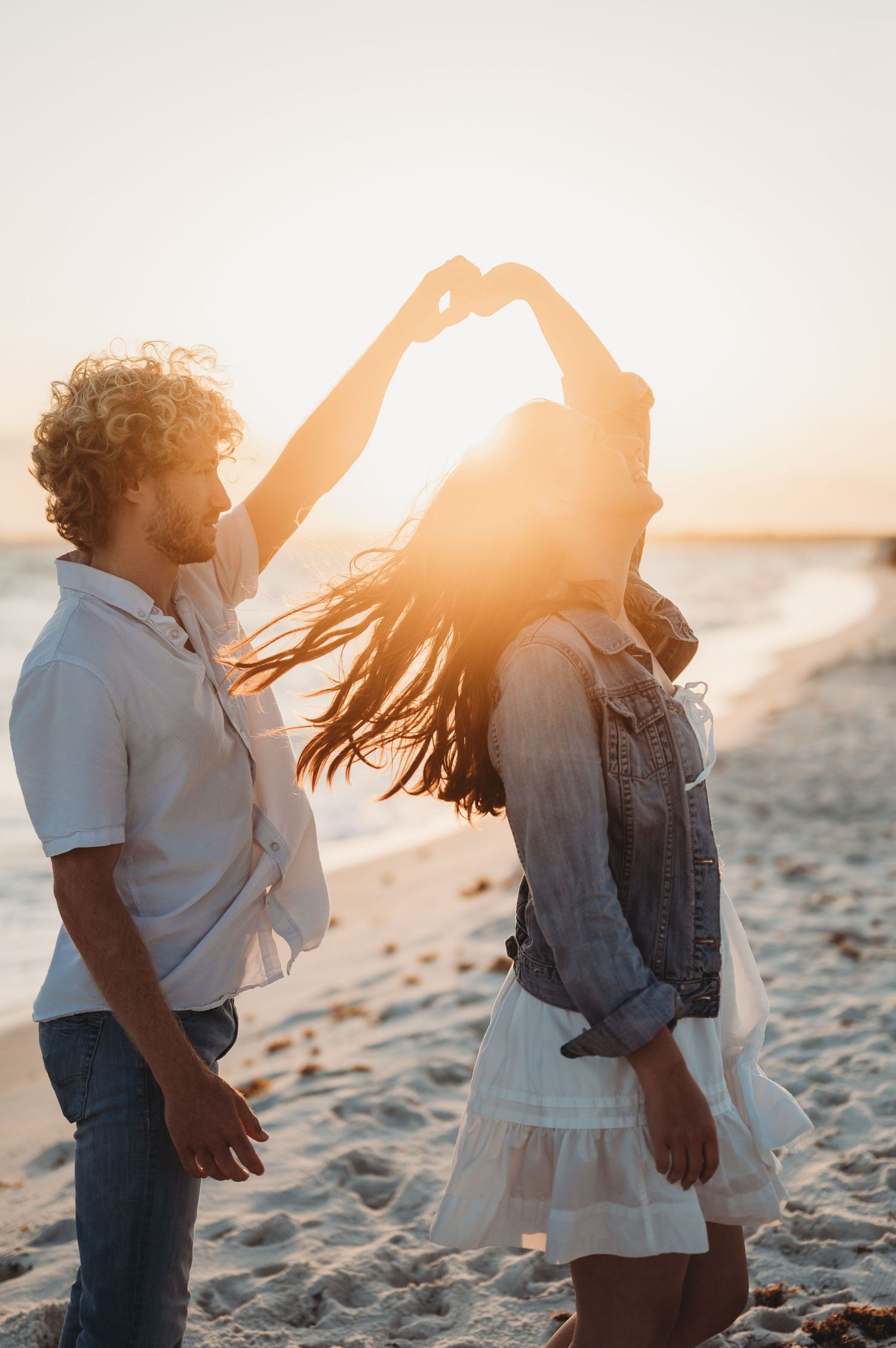 Couple dancing on beach at sunset, woman making heart shape with hands above her head.