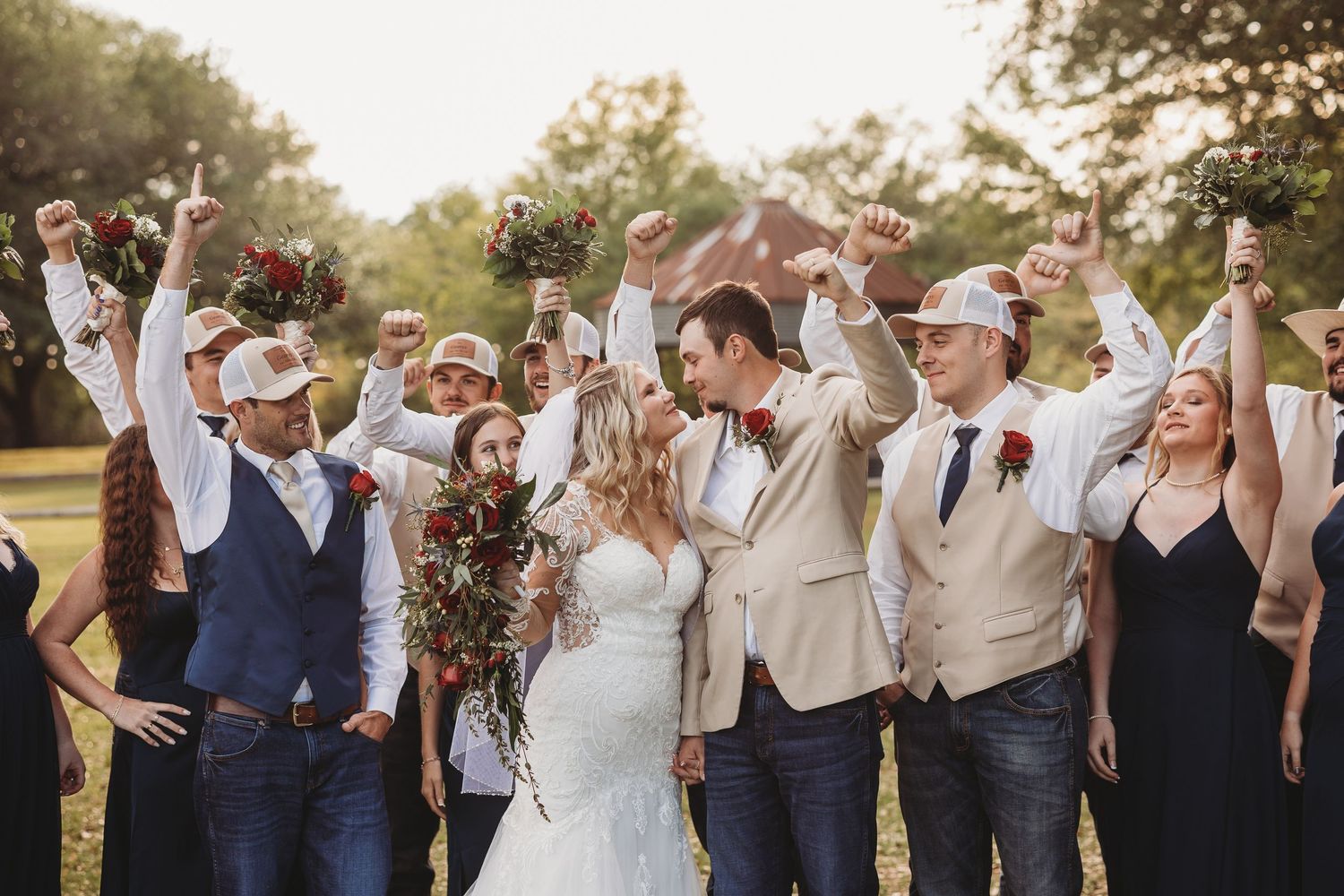 Wedding party celebrating outdoors with raised fists, bride and groom kissing, groomsmen in cowboy hats.