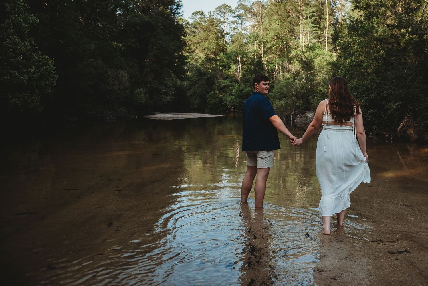 Romantic couple holding hands while wading through a tranquil forest river on a sunny day.