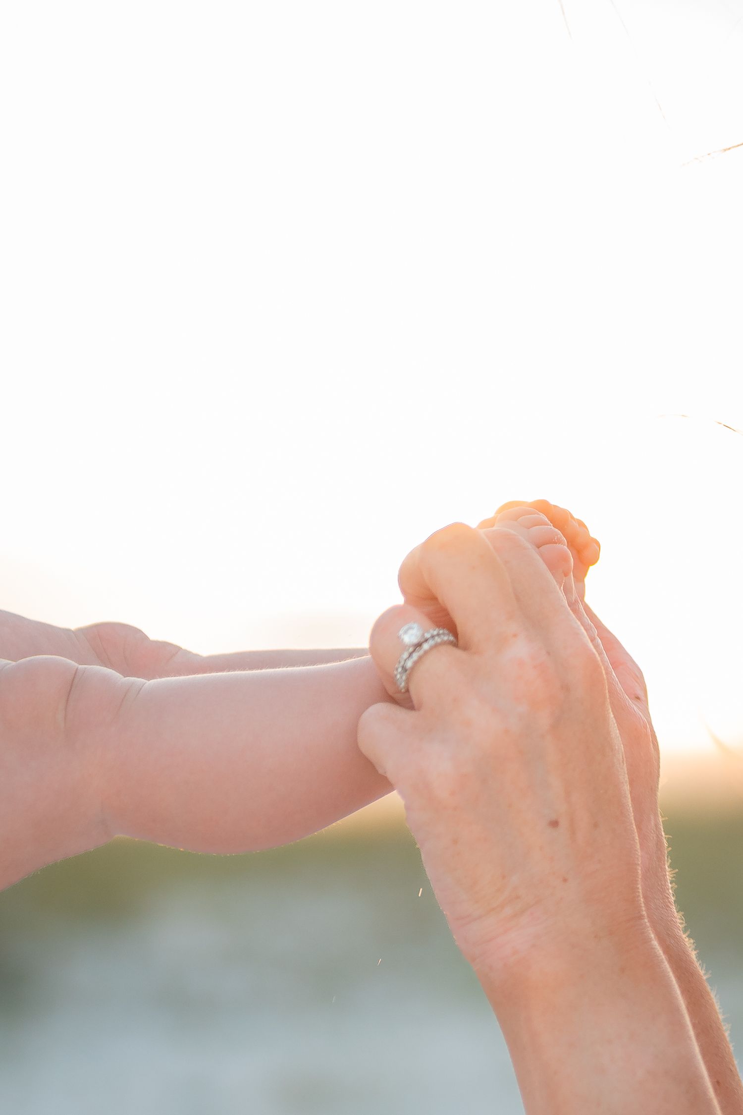 Adult hands gently holding a baby's tiny foot at sunset, with a diamond ring visible on one hand.