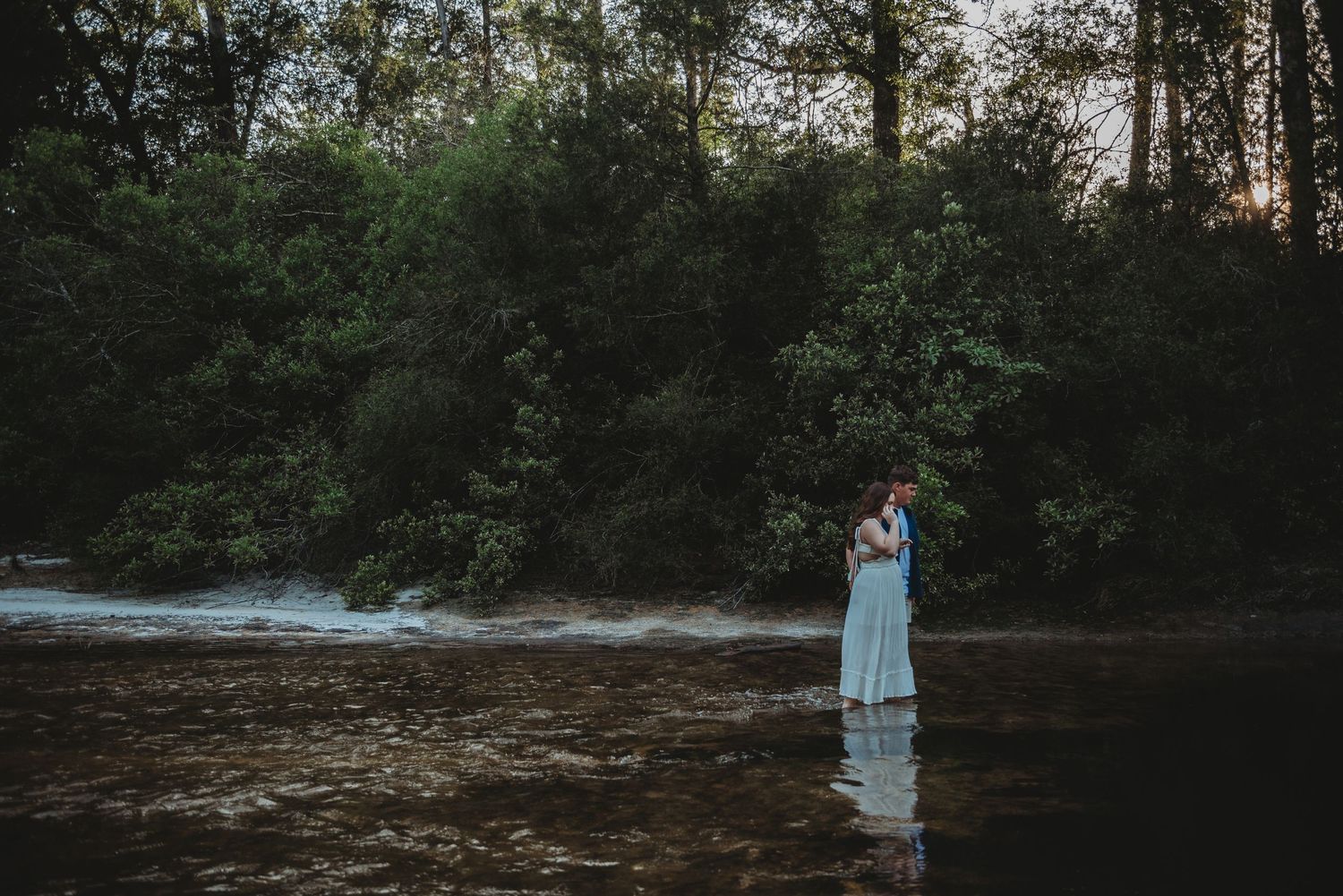 Couple embracing romantically while standing in a flowing river at dusk with lush forest backdrop.