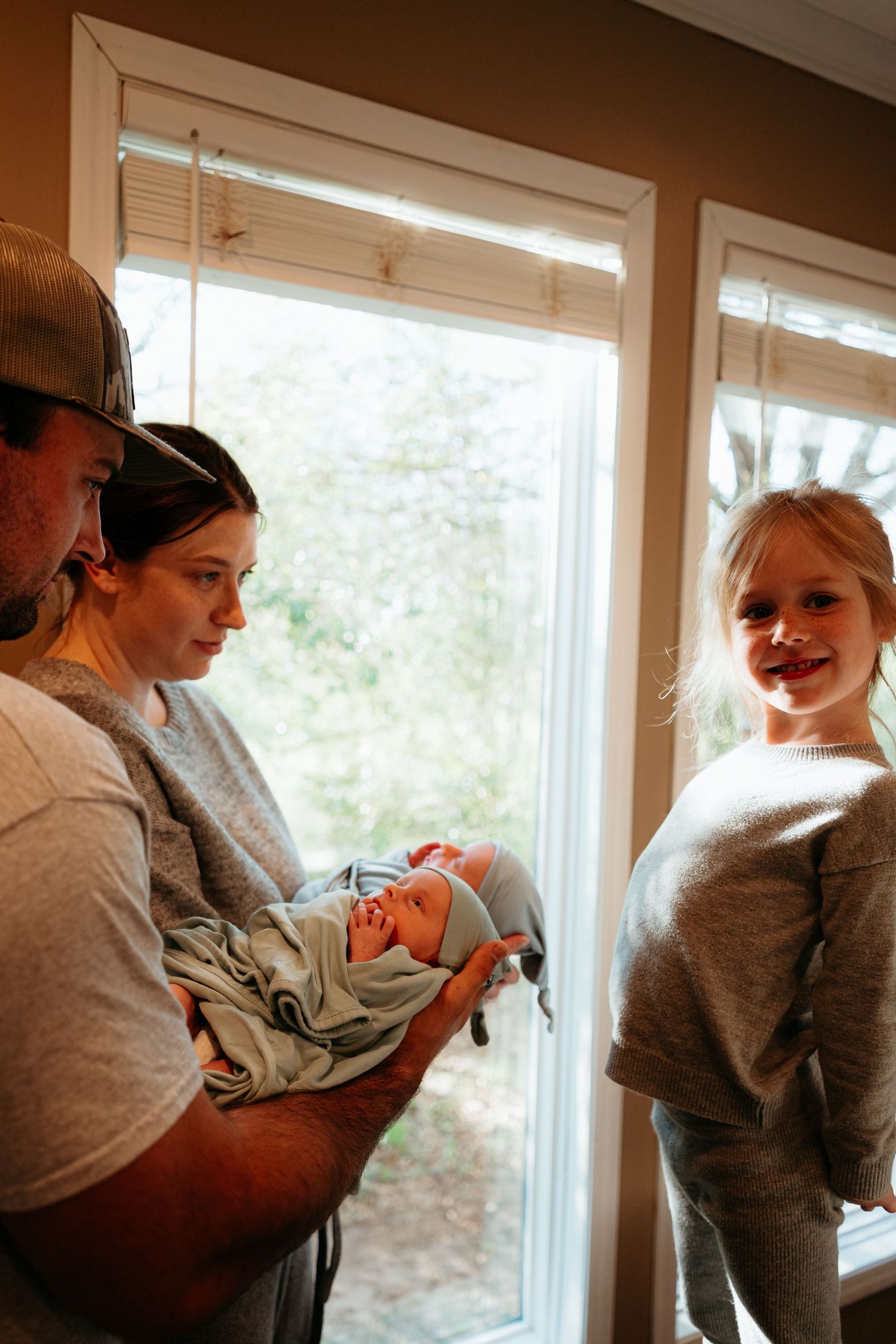 Young family with father in cap, mother holding newborn, and smiling toddler girl by window.