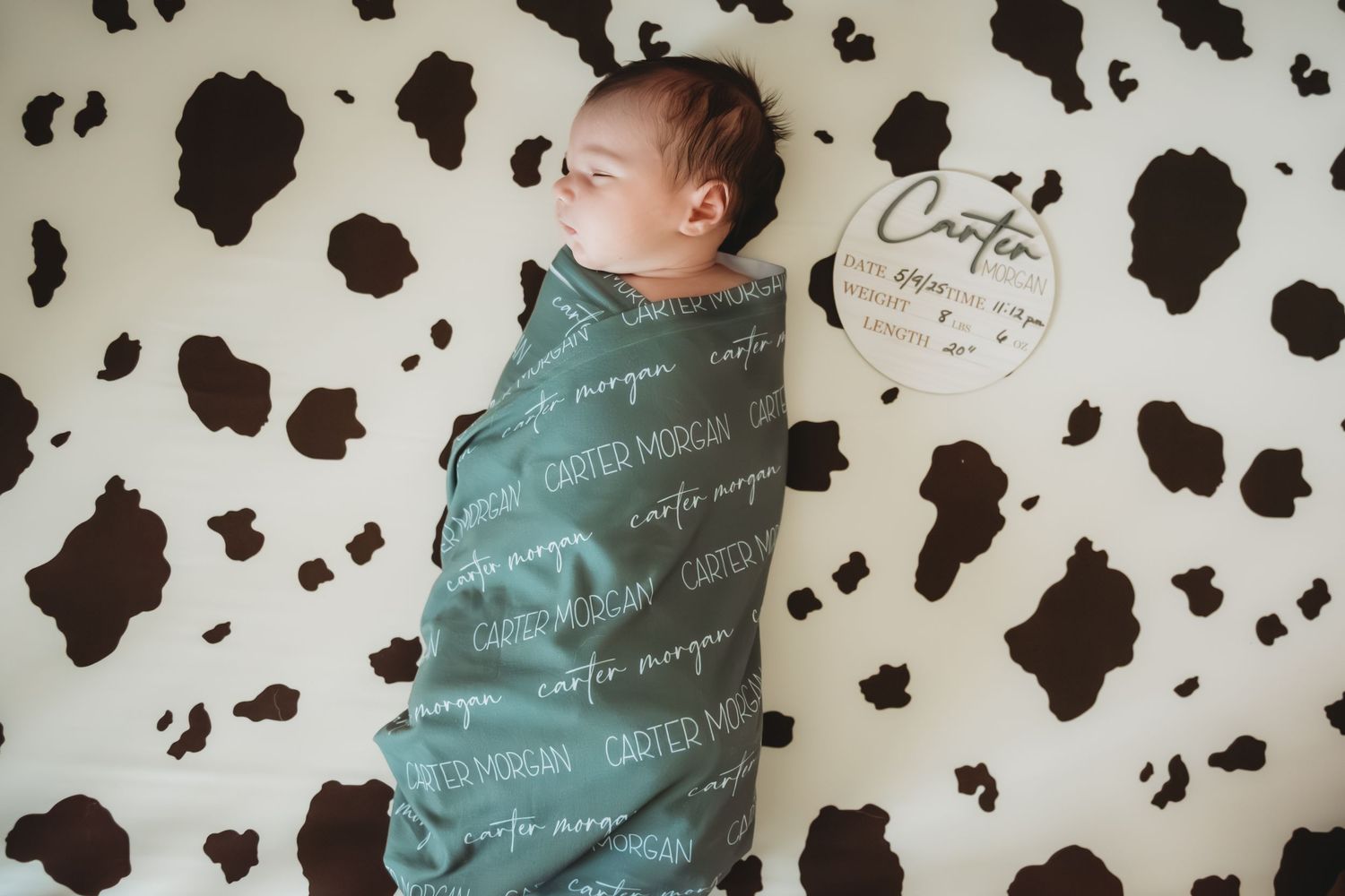 Sleeping newborn wrapped in teal name blanket on cow print sheet with wooden name sign.