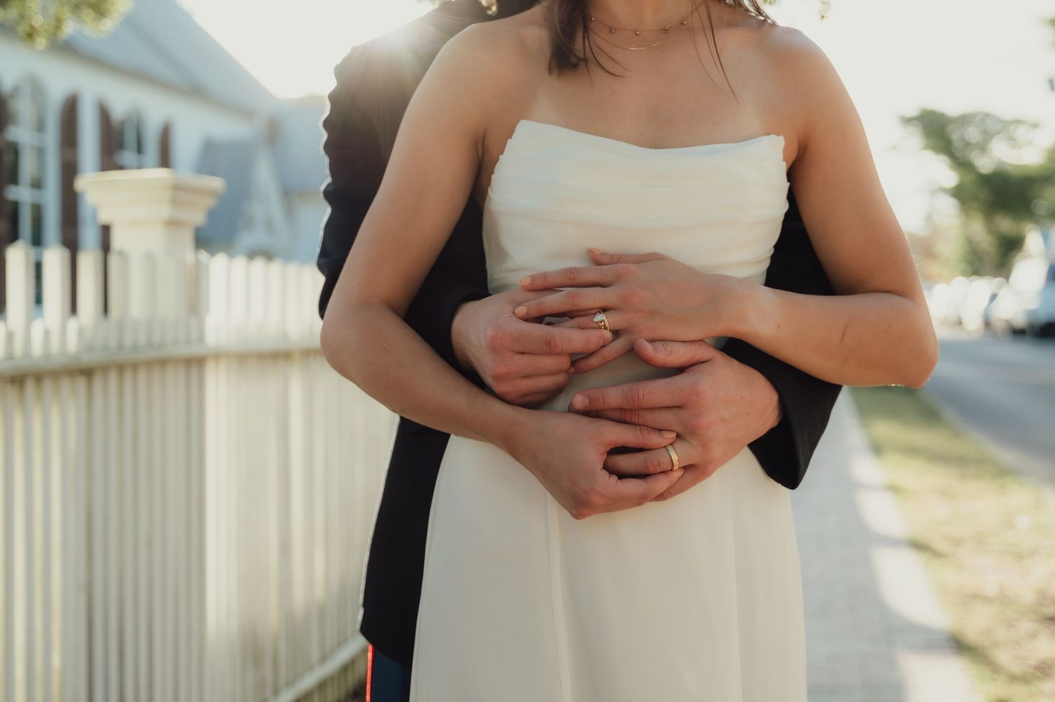 Couple embracing outdoors near white picket fence, bride in strapless white dress, groom in dark suit, wedding rings visible.