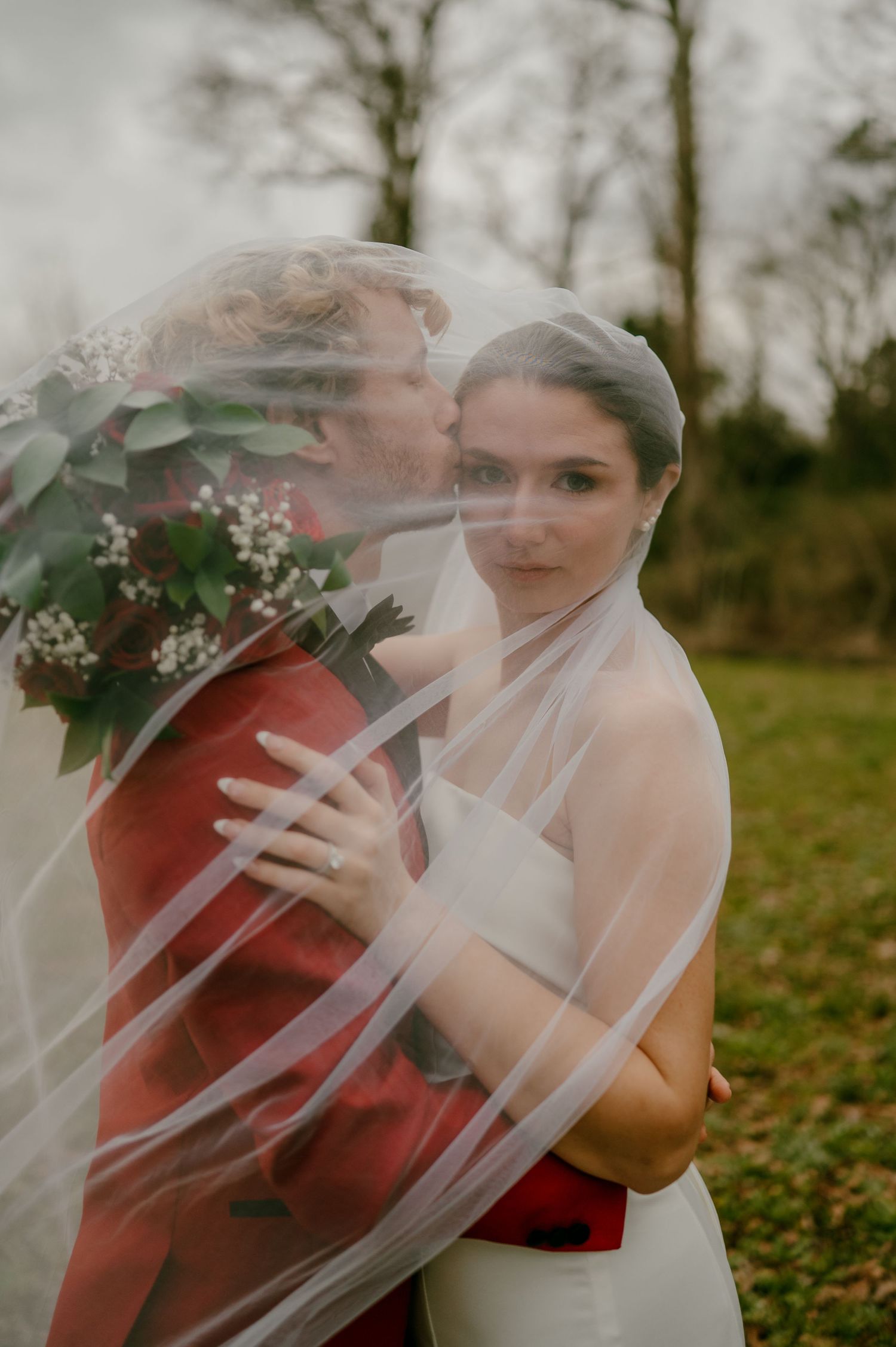 Bride and groom embrace under a flowing veil outdoors, groom in red suit kissing her cheek while she holds a bouquet.