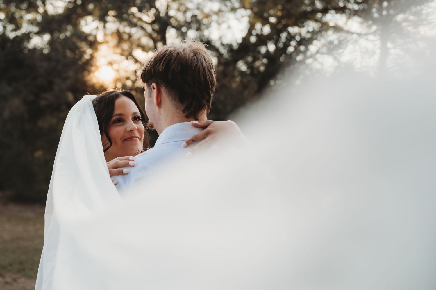 A bride and groom share a romantic embrace outdoors, wrapped in a white veil, with soft sunlight filtering through trees.