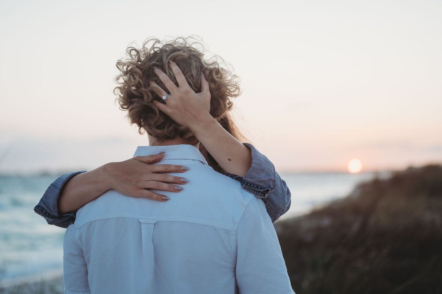 Couple embracing at sunset on a rocky beach, woman resting hand on man's shoulder with curly hair.