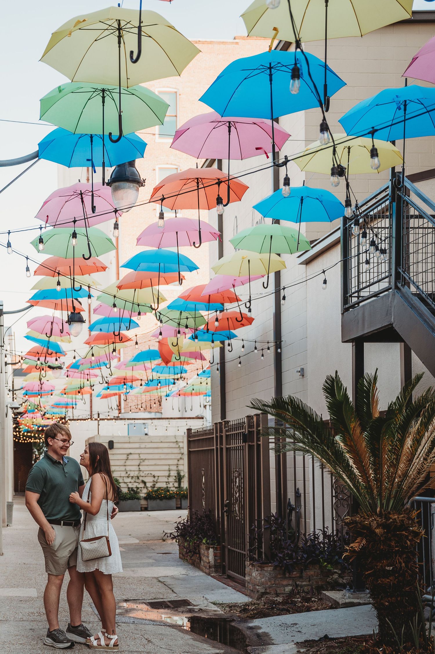 Couple standing together in colorful umbrella alley with multicolored umbrellas hanging overhead.