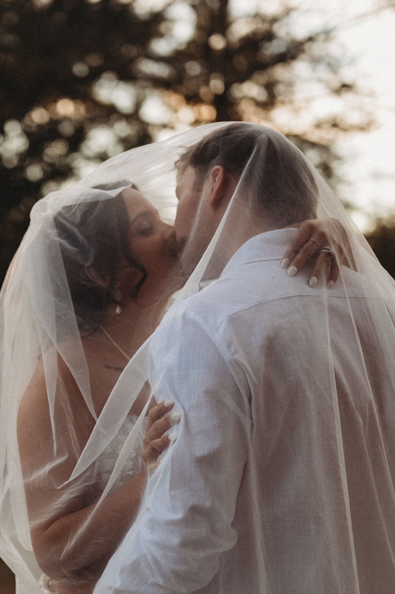 A bride and groom share a romantic kiss under a flowing white veil during their outdoor wedding ceremony.