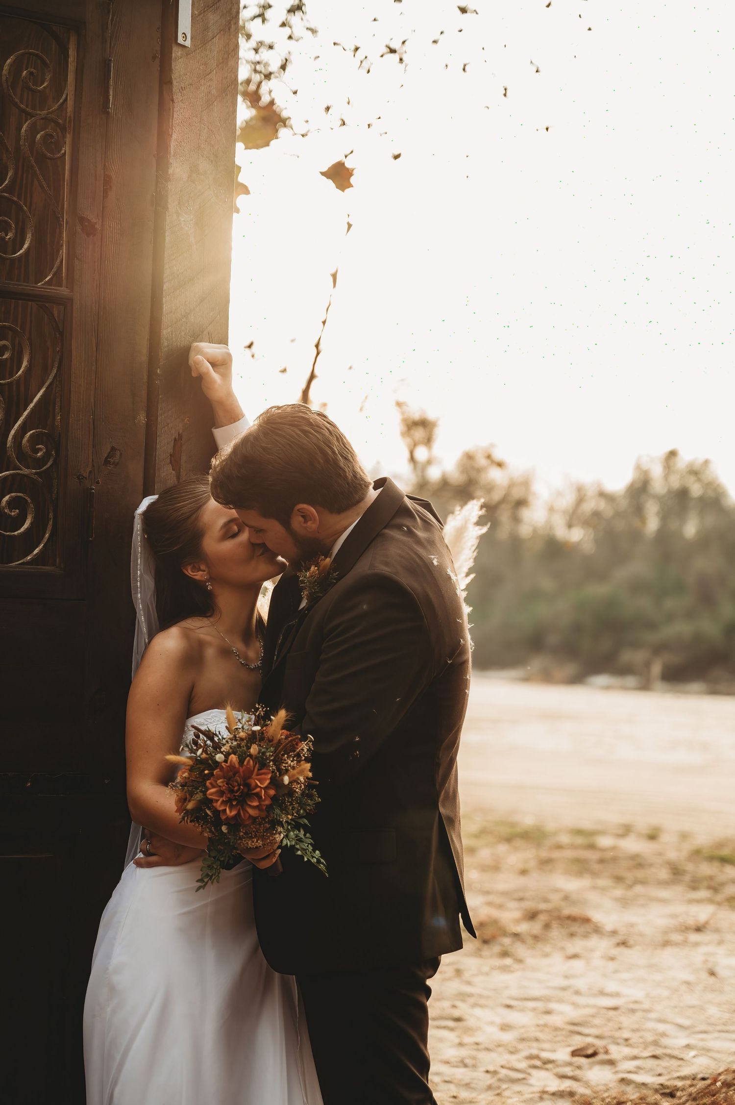 Bride and groom share a romantic kiss at golden hour beside an ornate wooden door, bride holding autumn bouquet.