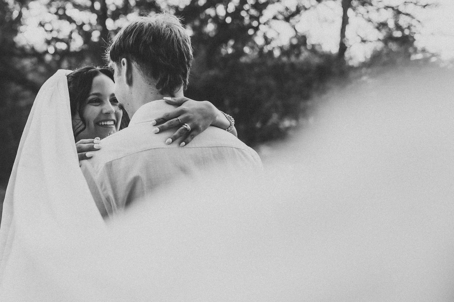 Black and white photo of a smiling bride embracing her groom outdoors on their wedding day.