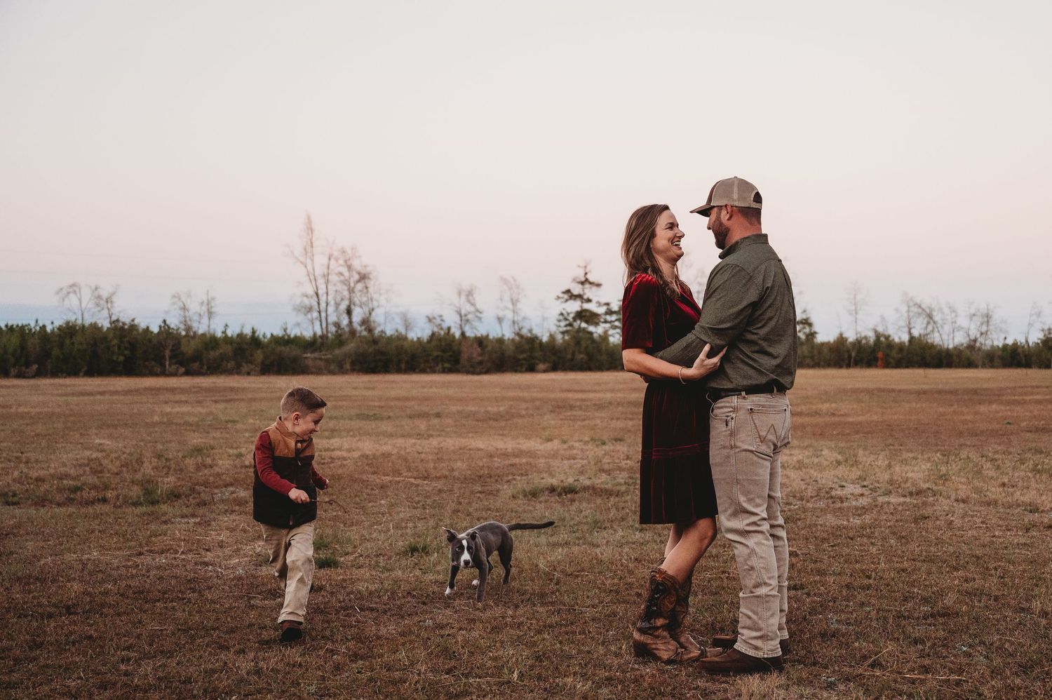 Family portrait at dusk in open field, couple embracing while child plays with dog in background.