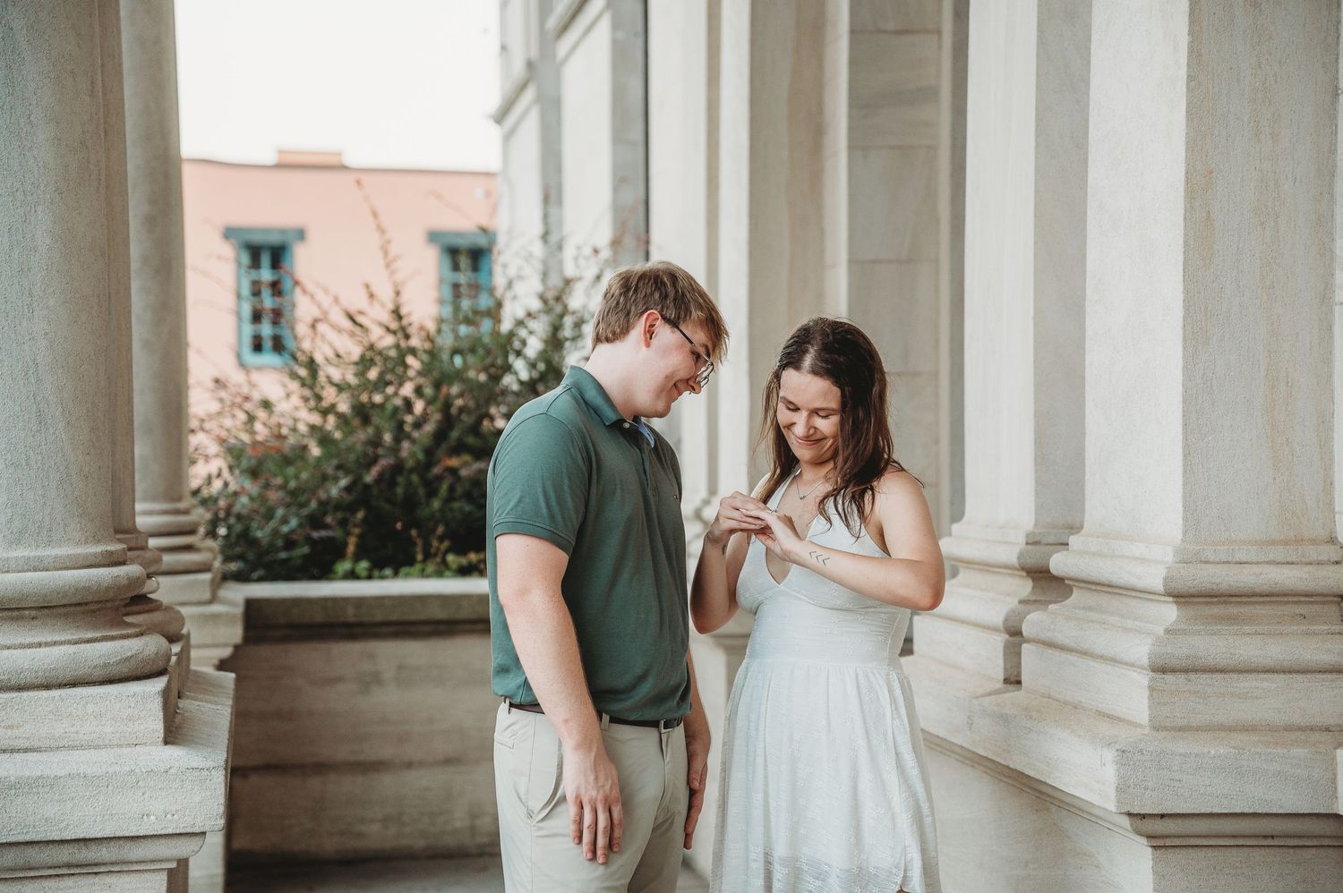 Engaged couple looking at a ring between elegant white columns, man in green polo and woman in white dress.
