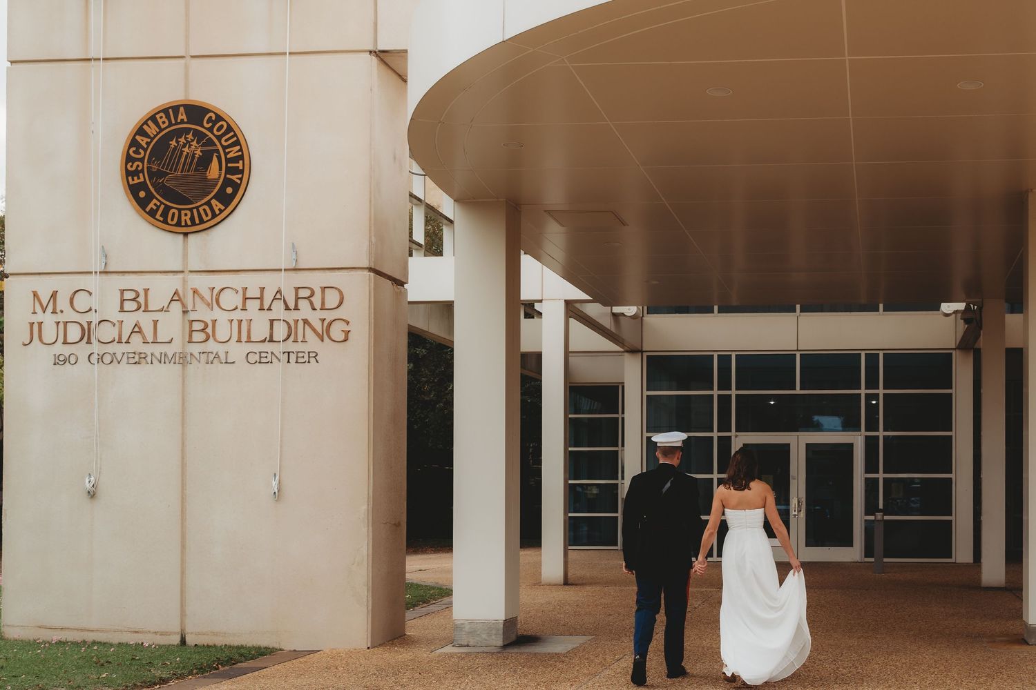 Couple walks hand-in-hand toward M.C. Blanchard Judicial Building entrance, bride in white gown, groom in dark attire.
