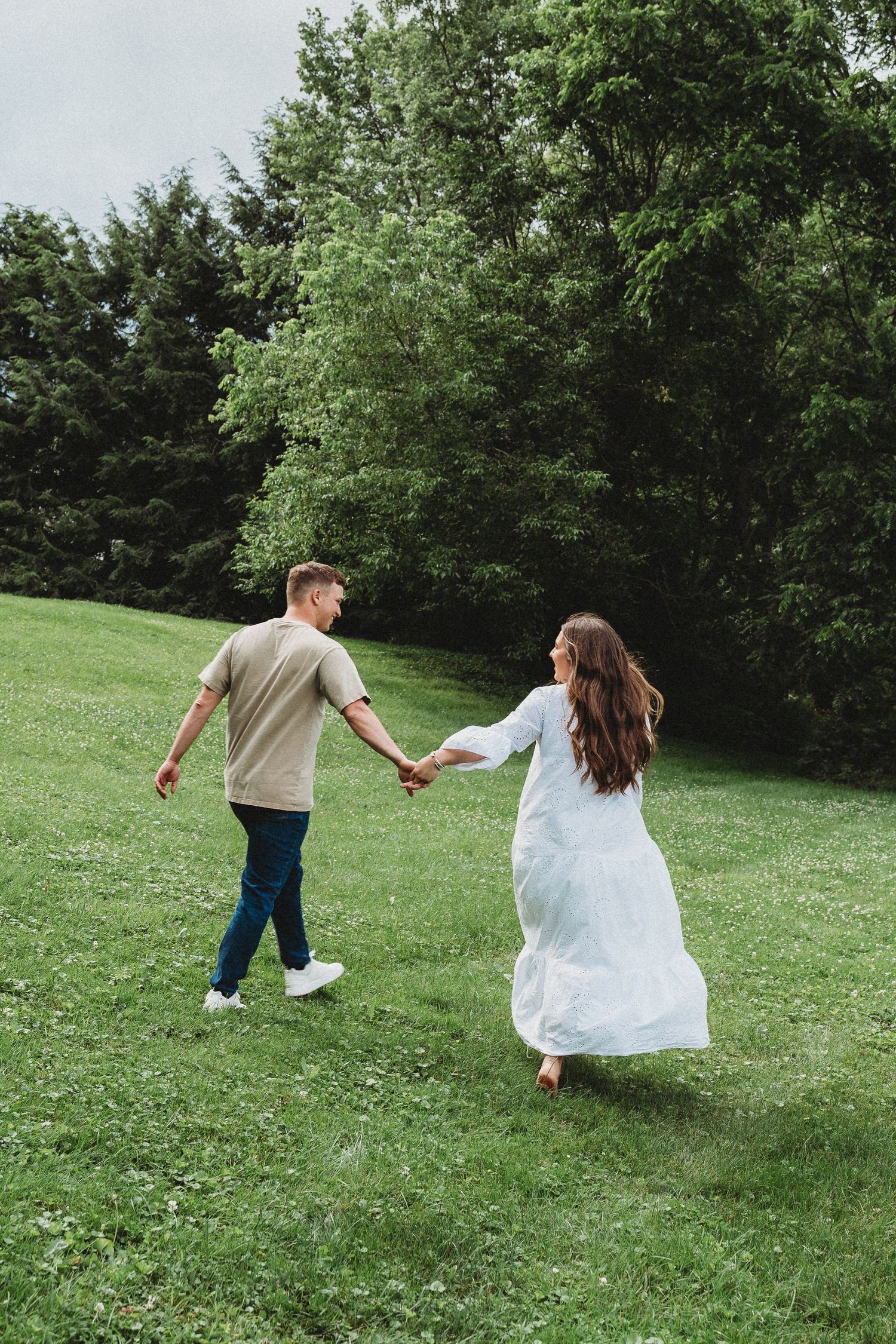 Couple holding hands walking on green grass lawn with lush trees in background, woman in white dress, man in casual outfit.