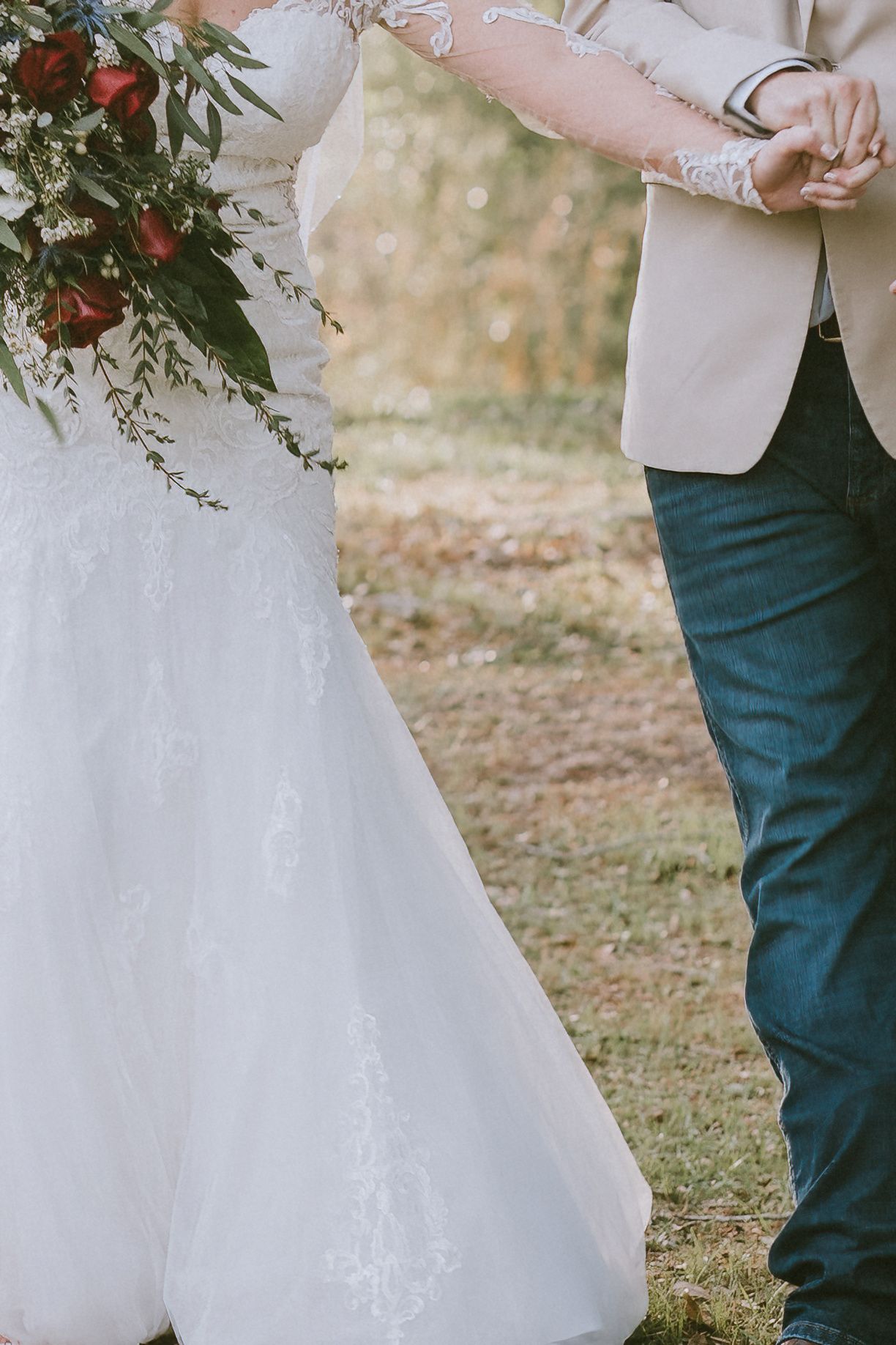 Bride in lace wedding gown holding red floral bouquet stands beside groom in beige blazer and jeans outdoors.