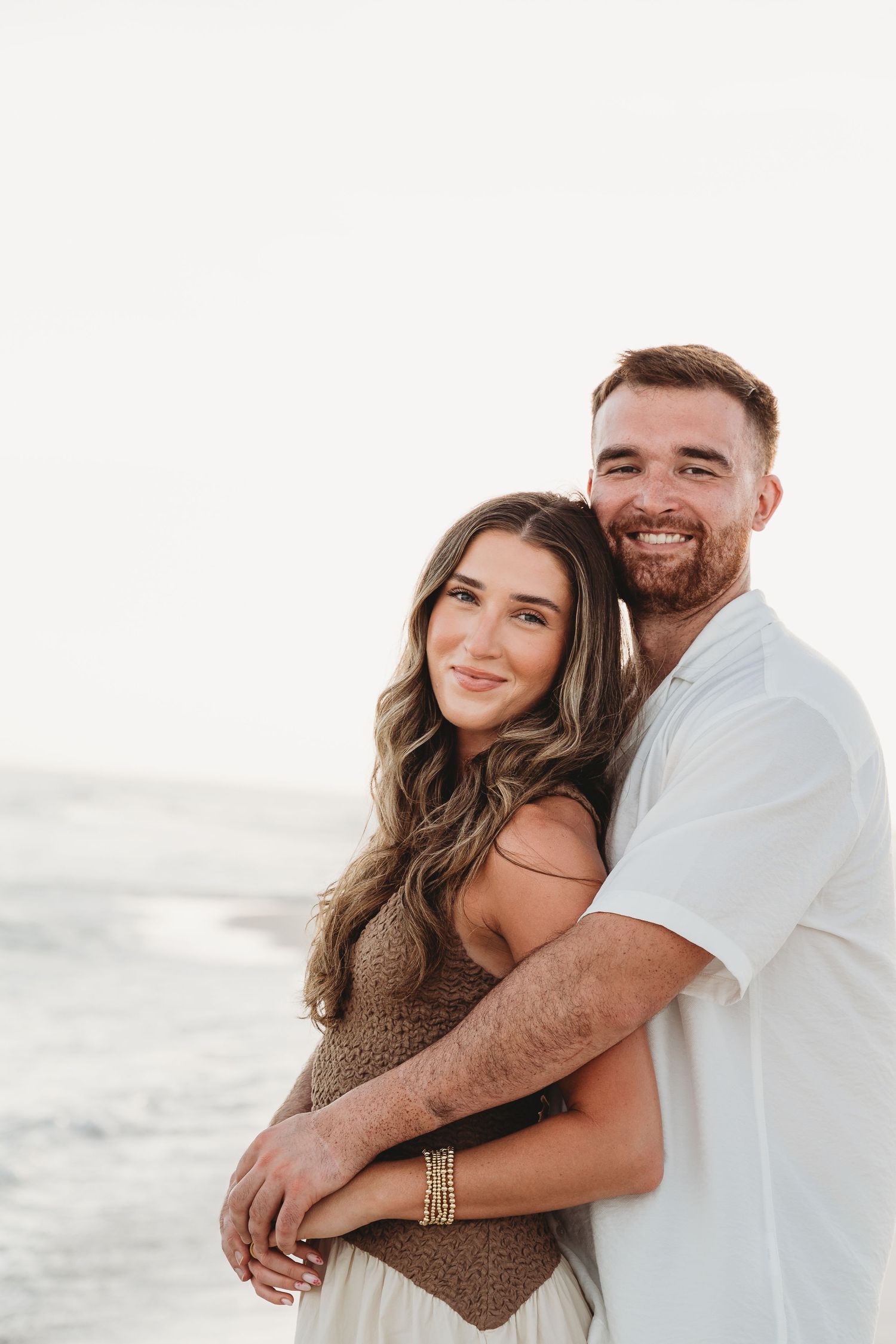 Smiling couple embracing on a beach at sunset, woman in brown top and man in white shirt.