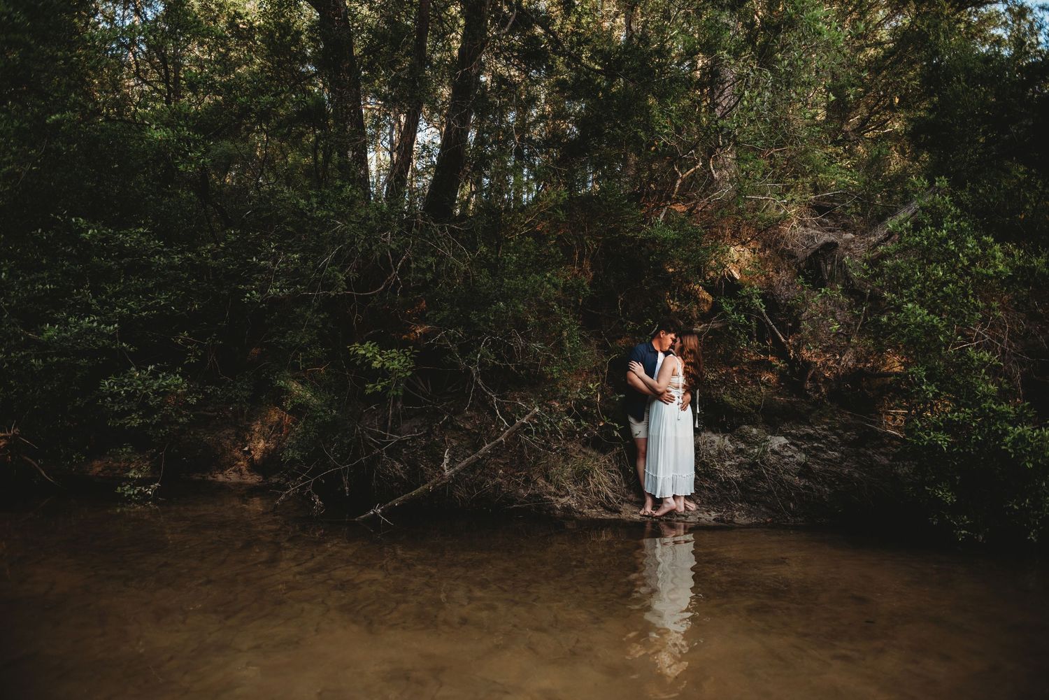 Couple kissing near fallen branches in a tranquil forest river setting with lush green trees.