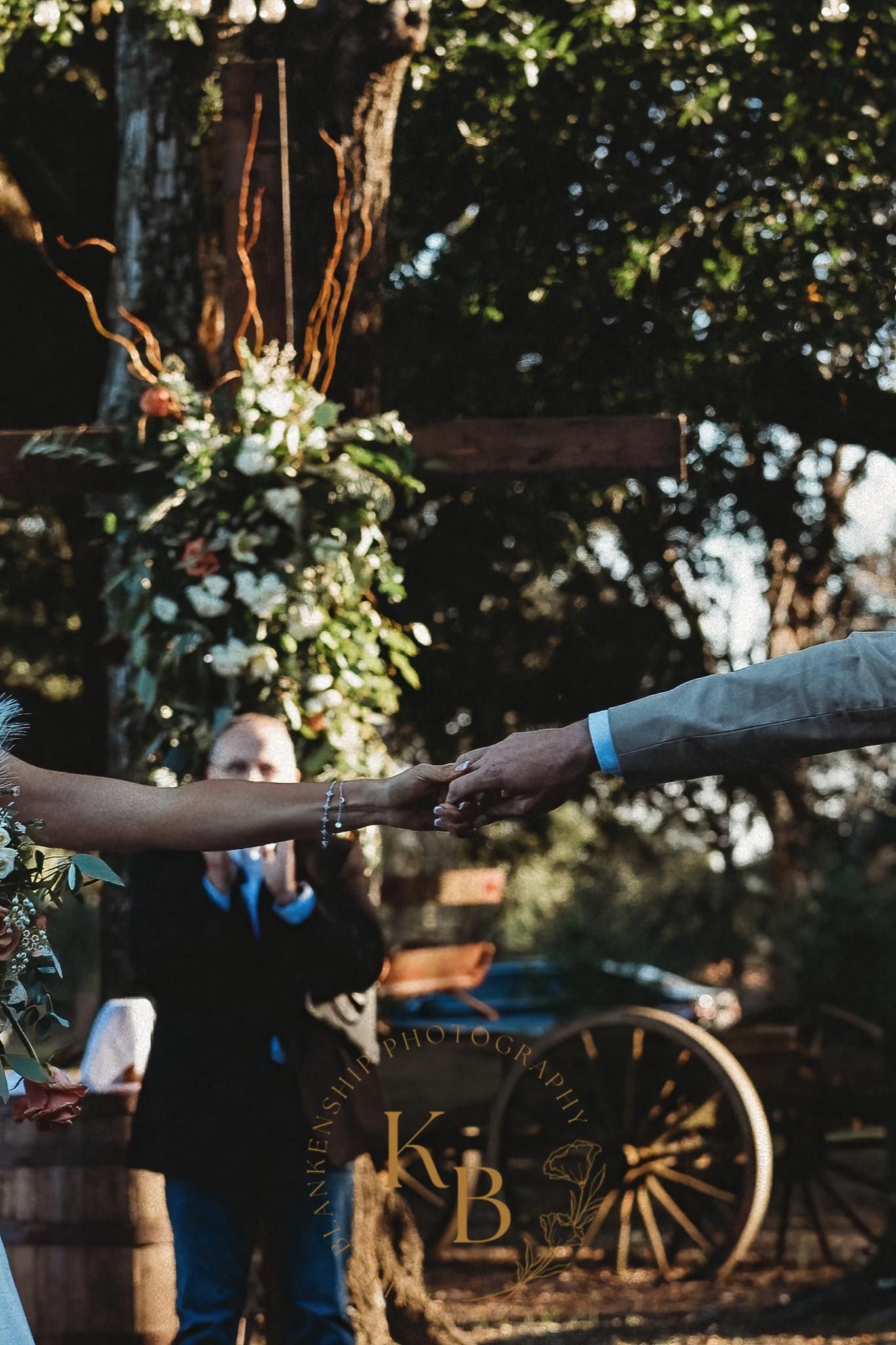 Couple exchanging vows outdoors, holding hands before officiant with rustic wooden cross backdrop.