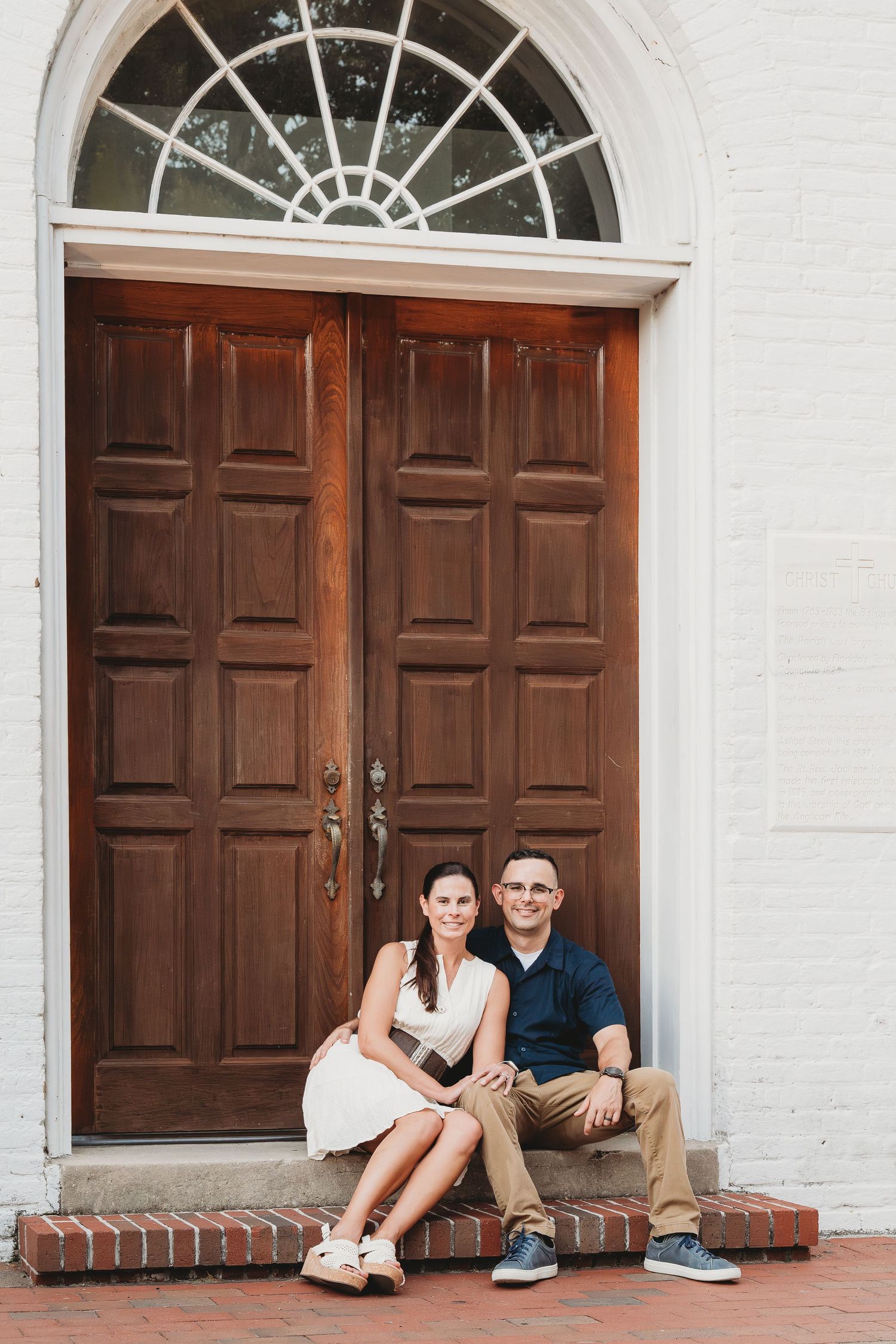 Smiling couple sitting together on brick steps in front of large wooden double doors.