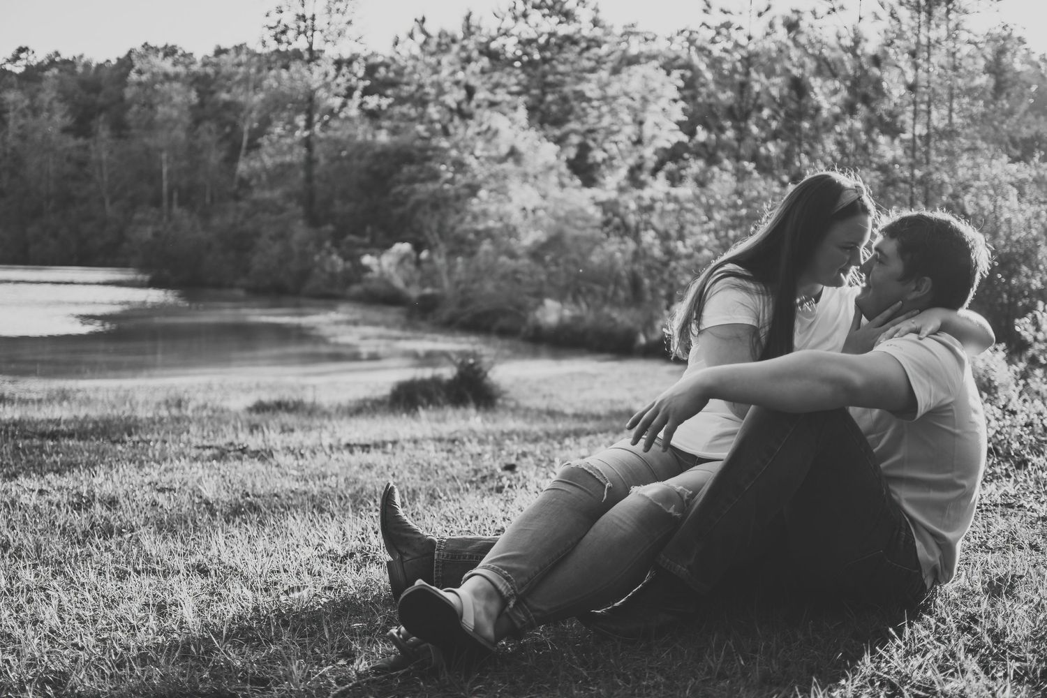 Black and white photo of couple sitting together on grass near a lake, sharing a quiet embrace.