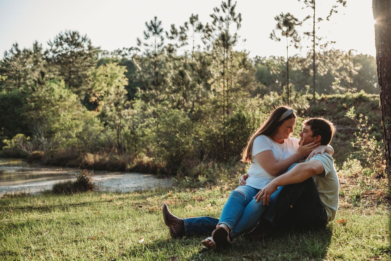 Romantic couple sitting close together on a grassy hillside near a pond in warm afternoon sunlight.